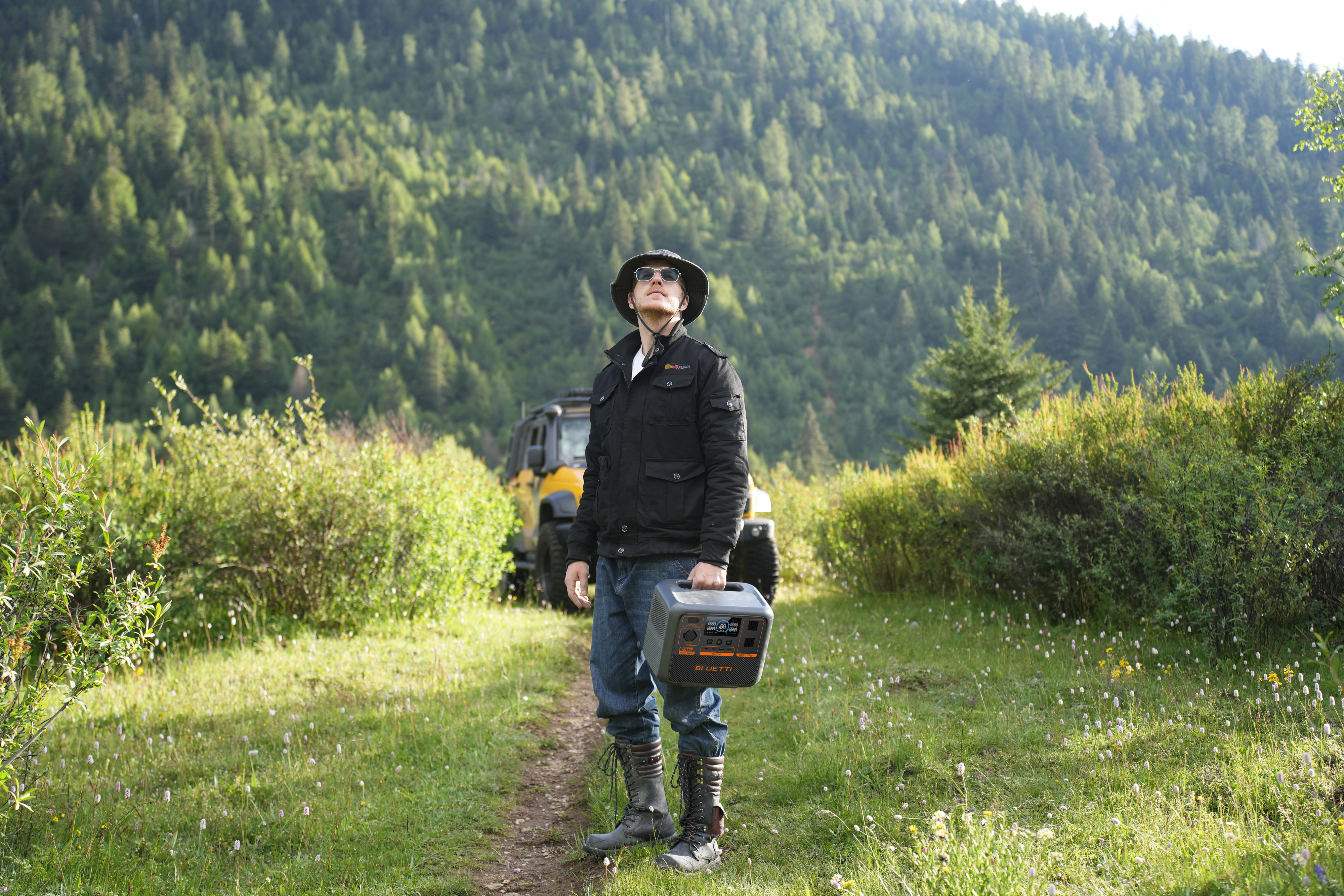 Mężczyzna trzymający elektrownię na polu, w kapeluszu, z pojazdem w tle.