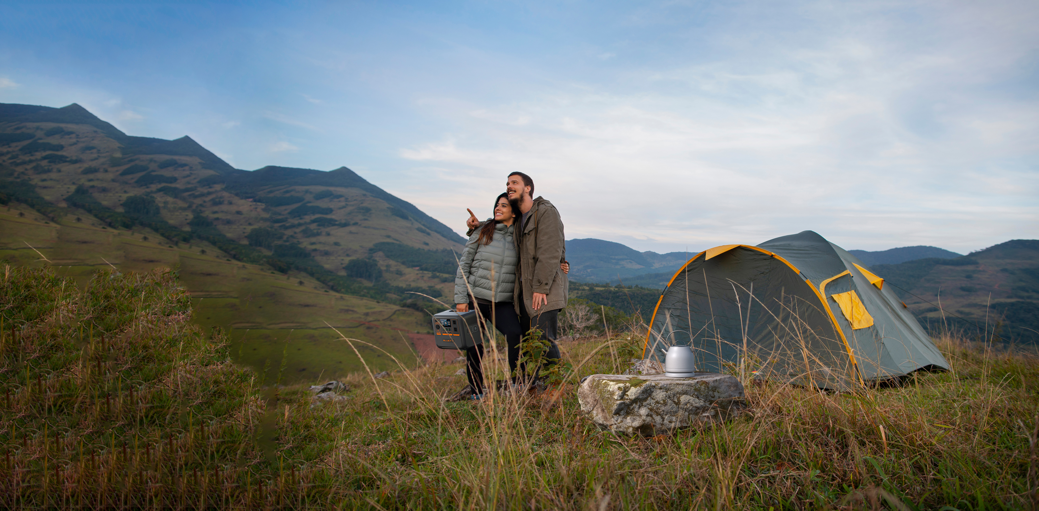 Para na łonie natury, wskazująca w górę. Namiot i sprzęt kempingowy w tle, widać góry i niebo.