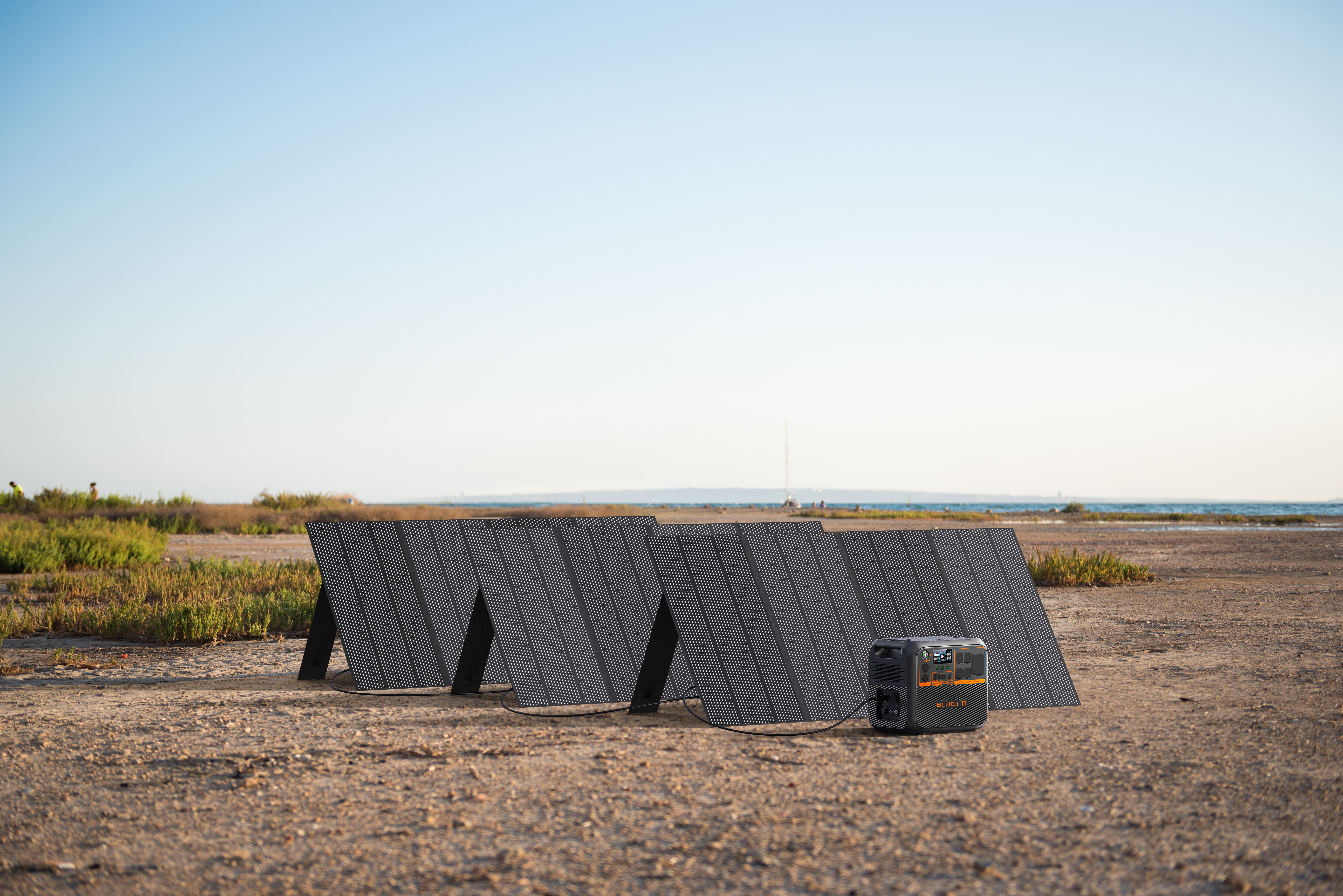 Panele słoneczne ustawione na polu. W tle stacja zasilania i widok na morze.