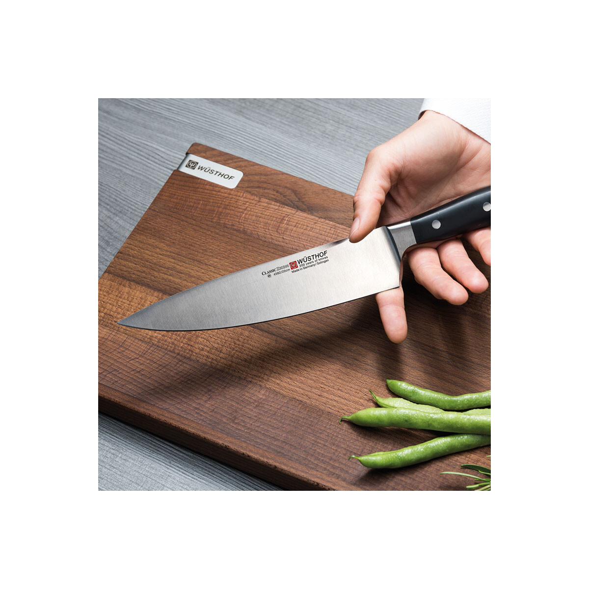 Hand hält Küchenmesser auf Holzschneidebrett mit Bohnen. Das Messer ist scharf.