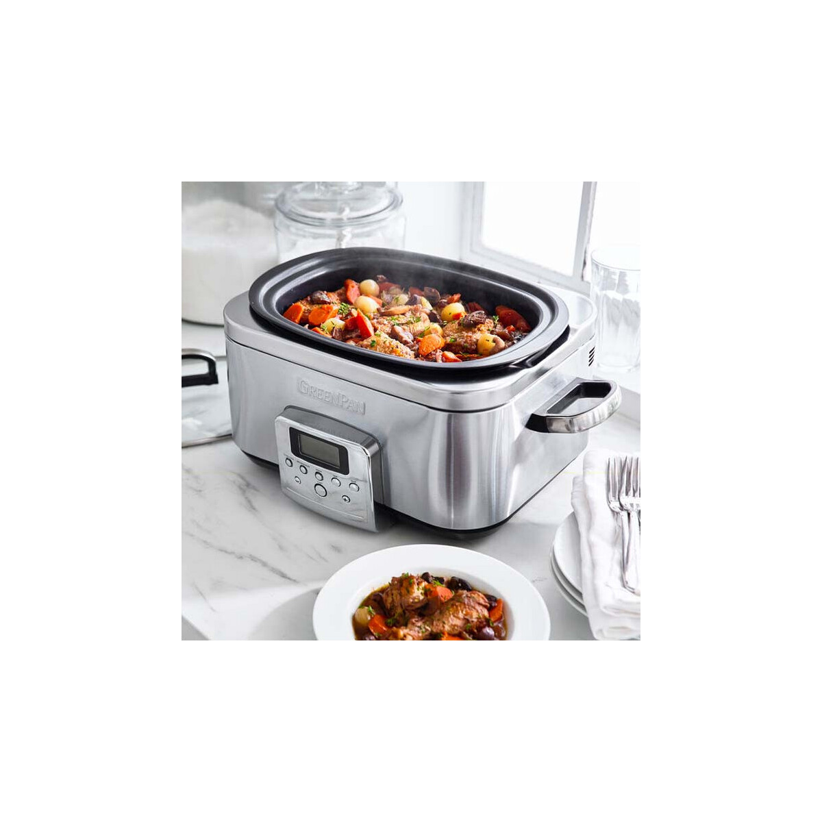 Silberner Slow Cooker mit Essen. Teller mit Essen davor. Glasbehälter und Fenster im Hintergrund.