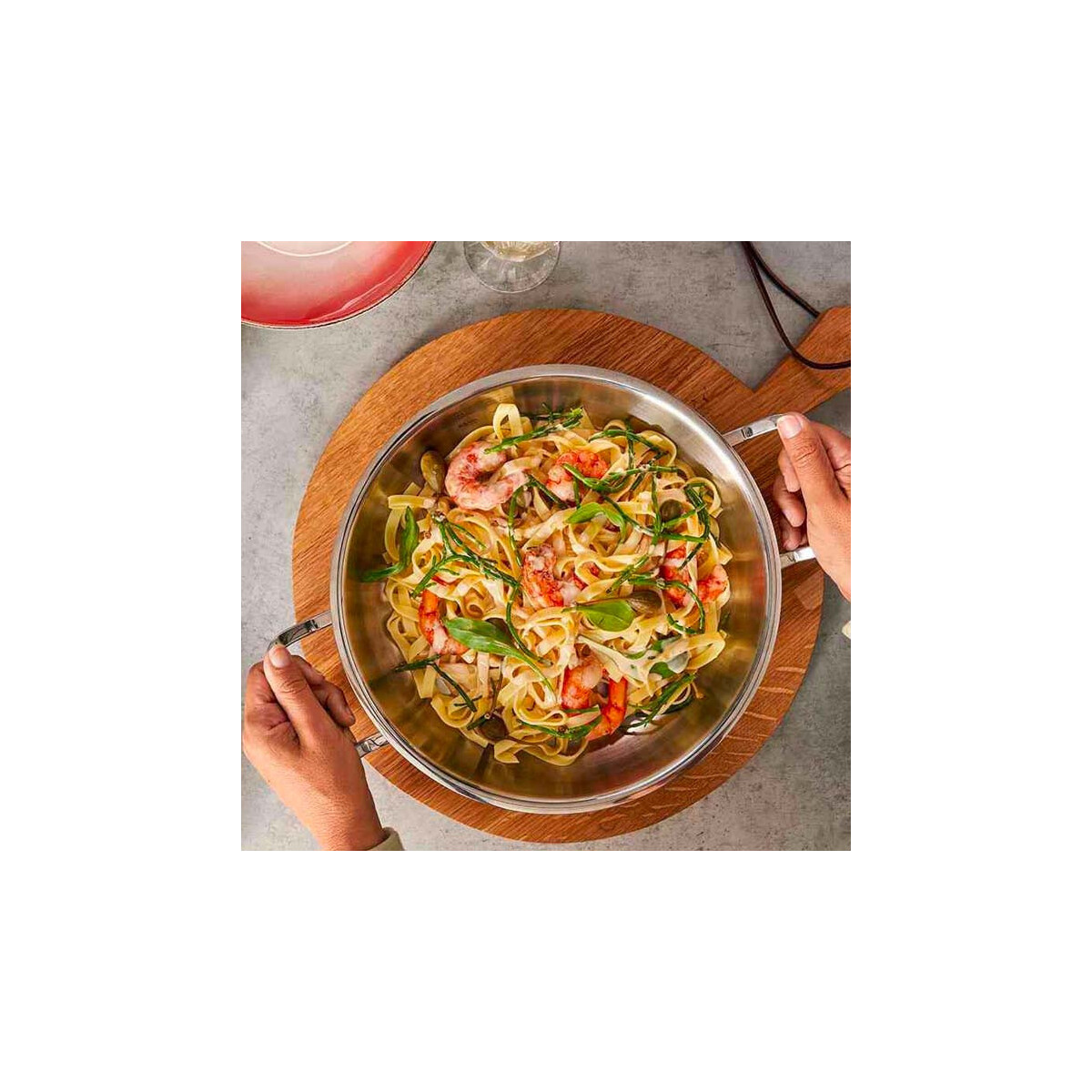 Pasta con gamberi in una padella d'acciaio su un tagliere di legno, tenuta da due mani.
