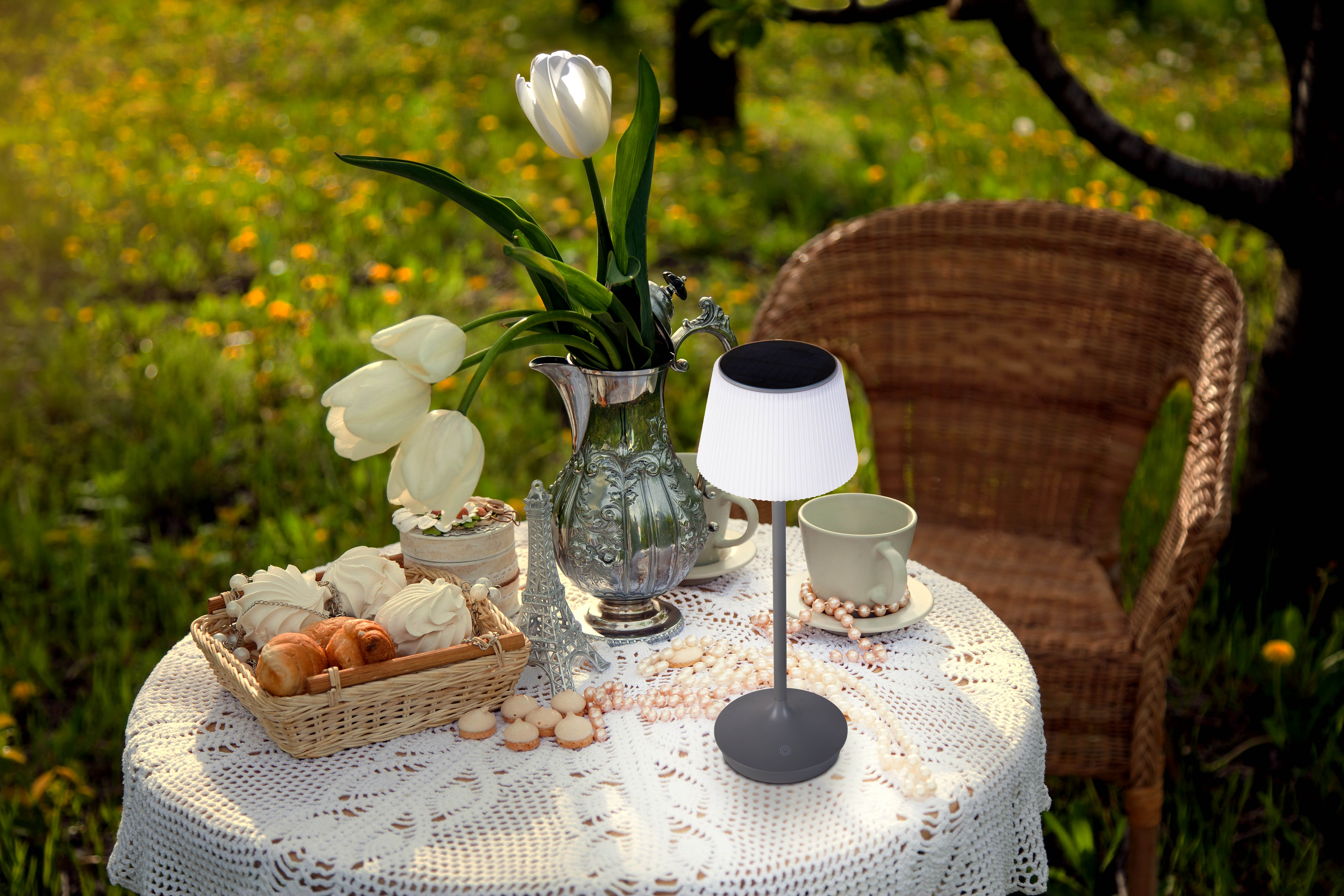 Szene im Freien mit einem Tisch für eine Mahlzeit. Enthält Blumen, Lampe und Gebäck. Korbstuhl und grünes Gras.