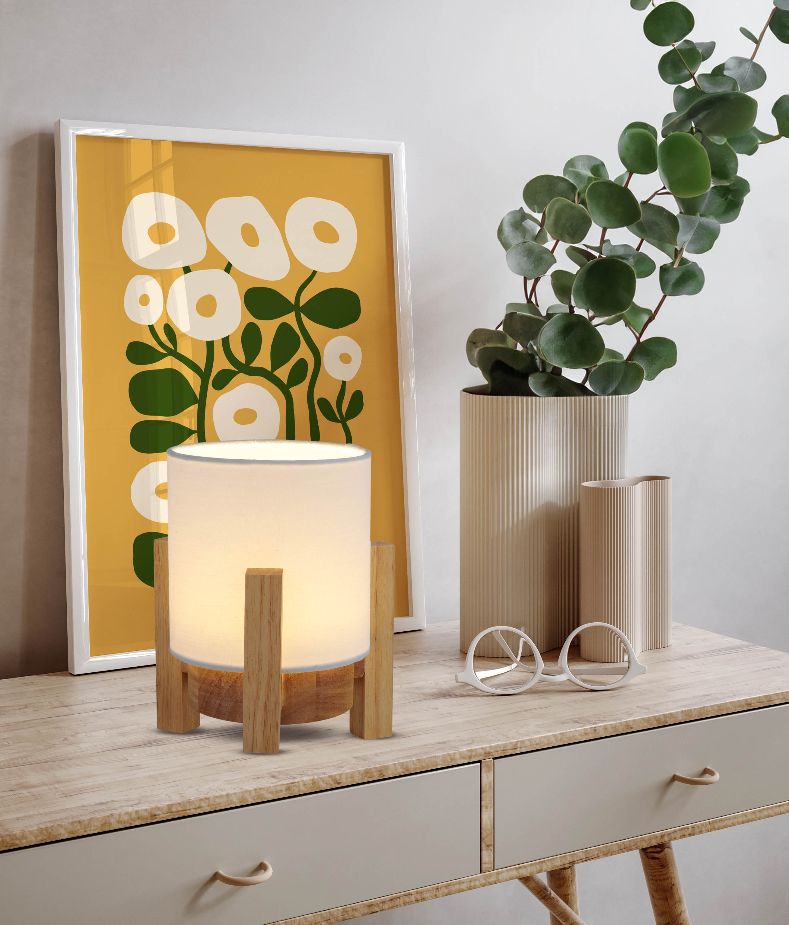 Eine Holzlampe mit weißem Schirm steht auf einem Tisch. Gelbe Kunst und Pflanze dahinter.
