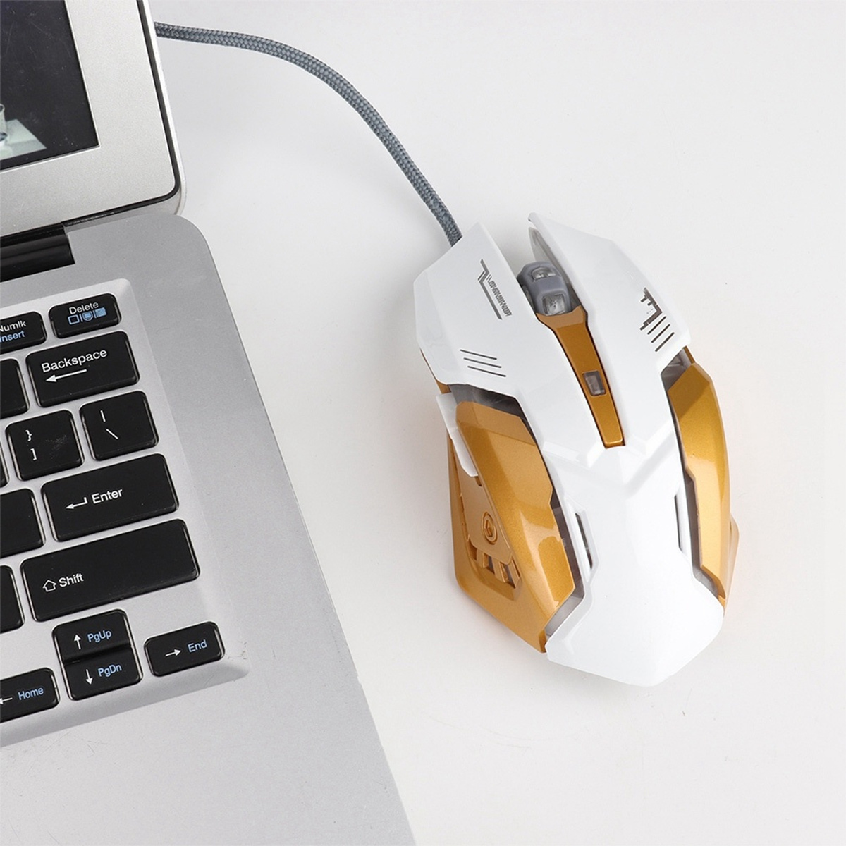 Eine weiße und goldene Gaming-Maus neben einem Laptop. Ein graues Kabel verbindet die Maus.