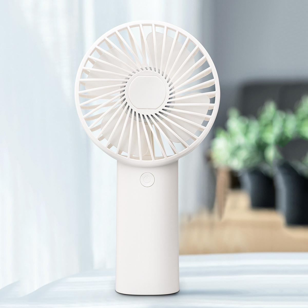 Ein weißer Handventilator steht auf einem Tisch. Im Hintergrund sind ein Fenster und grüne Pflanzen.