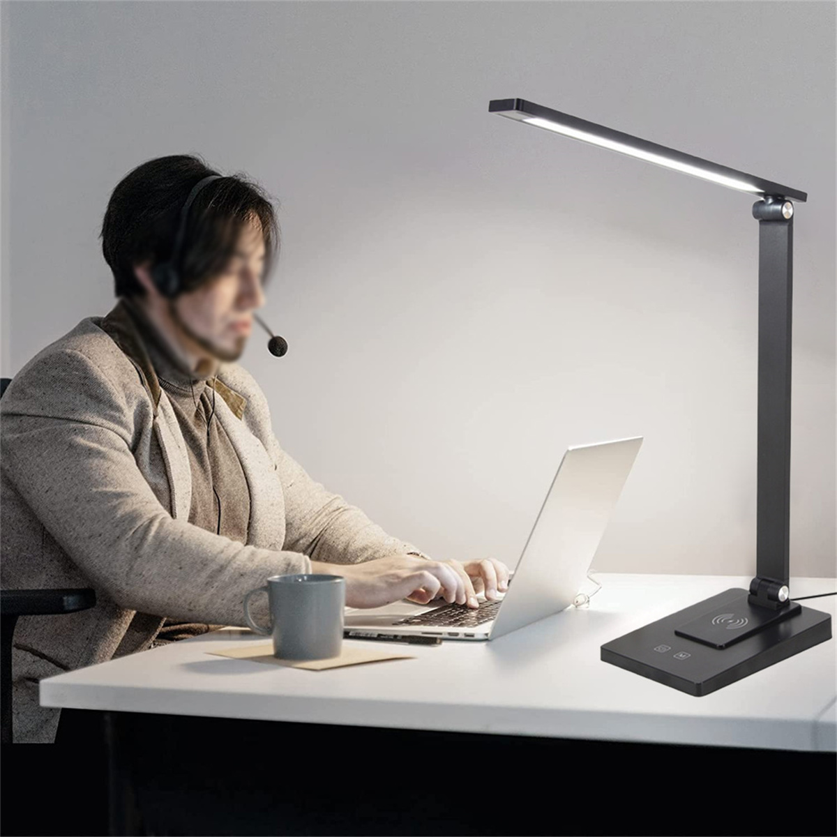 Ein Mann mit Headset arbeitet an einem Laptop unter einer schwarzen Schreibtischlampe. Die Lampe hat einen verstellbaren Arm und einen Sockel.