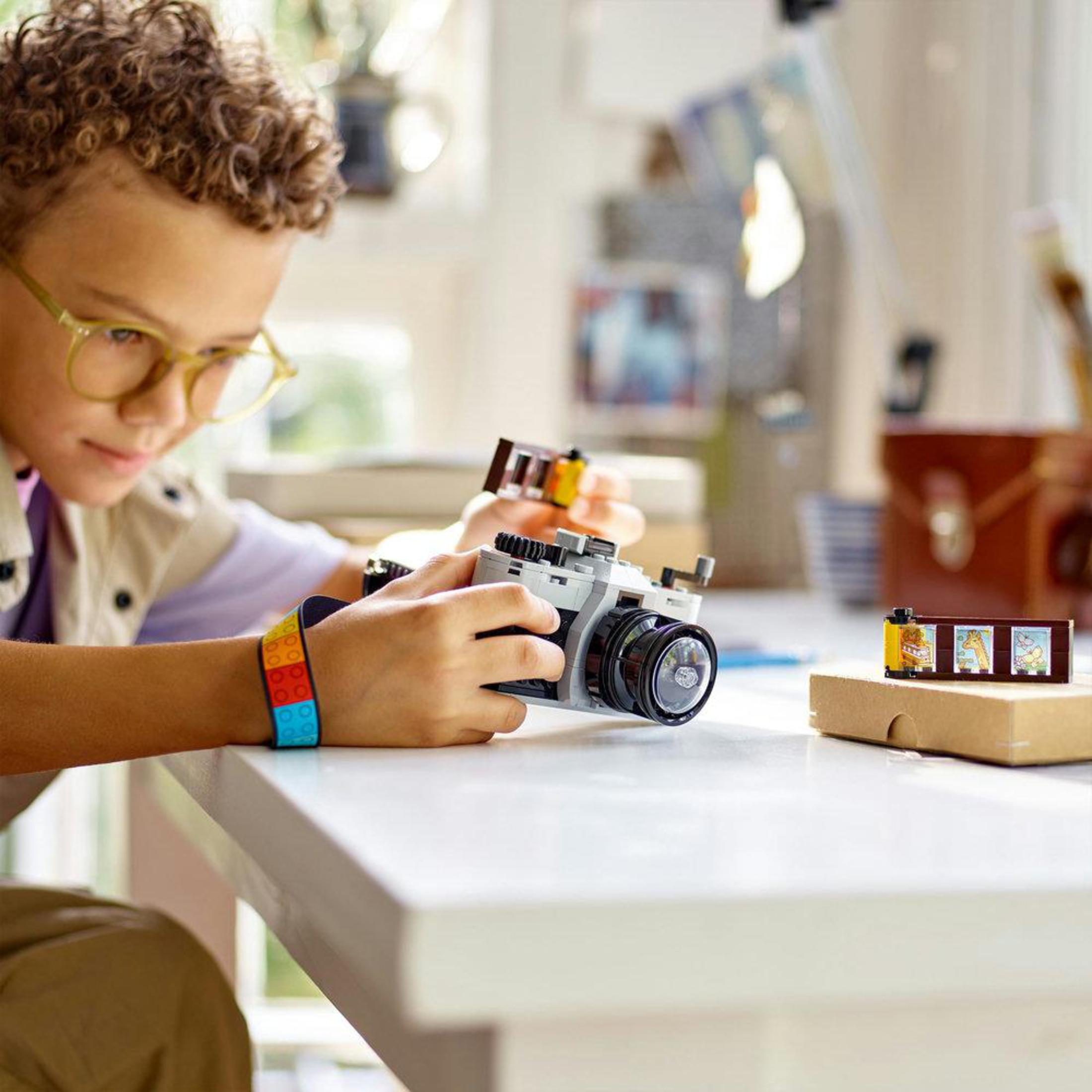 Un niño sosteniendo una cámara de juguete. Usa gafas y una pulsera colorida. Mesa blanca en la escena.
