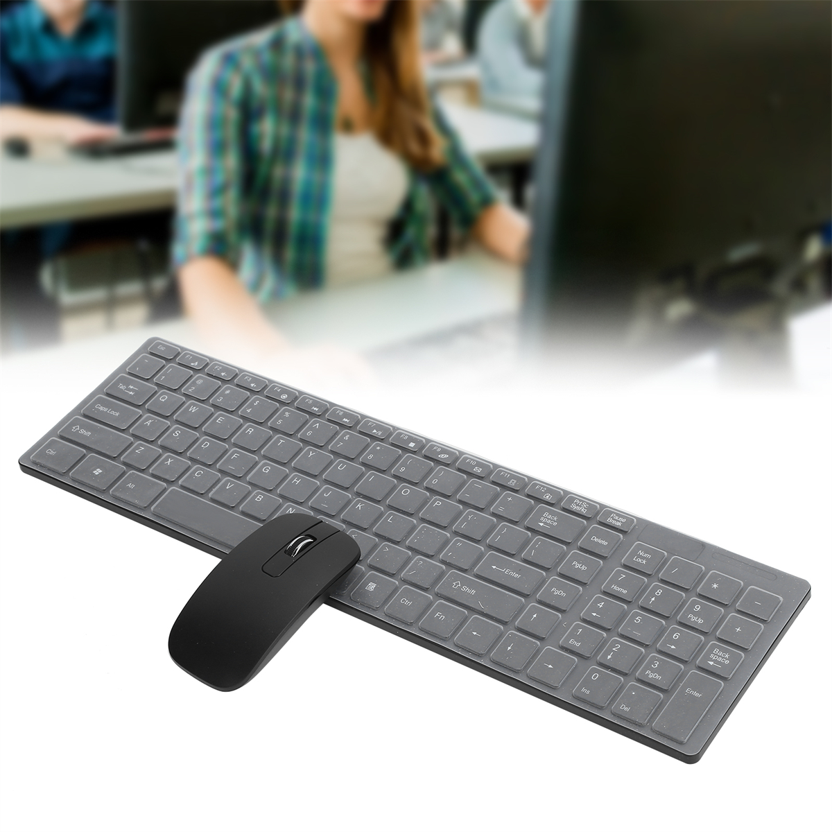 Eine graue Tastatur und Maus auf weißer Oberfläche, verschwommener Hintergrund einer Person.