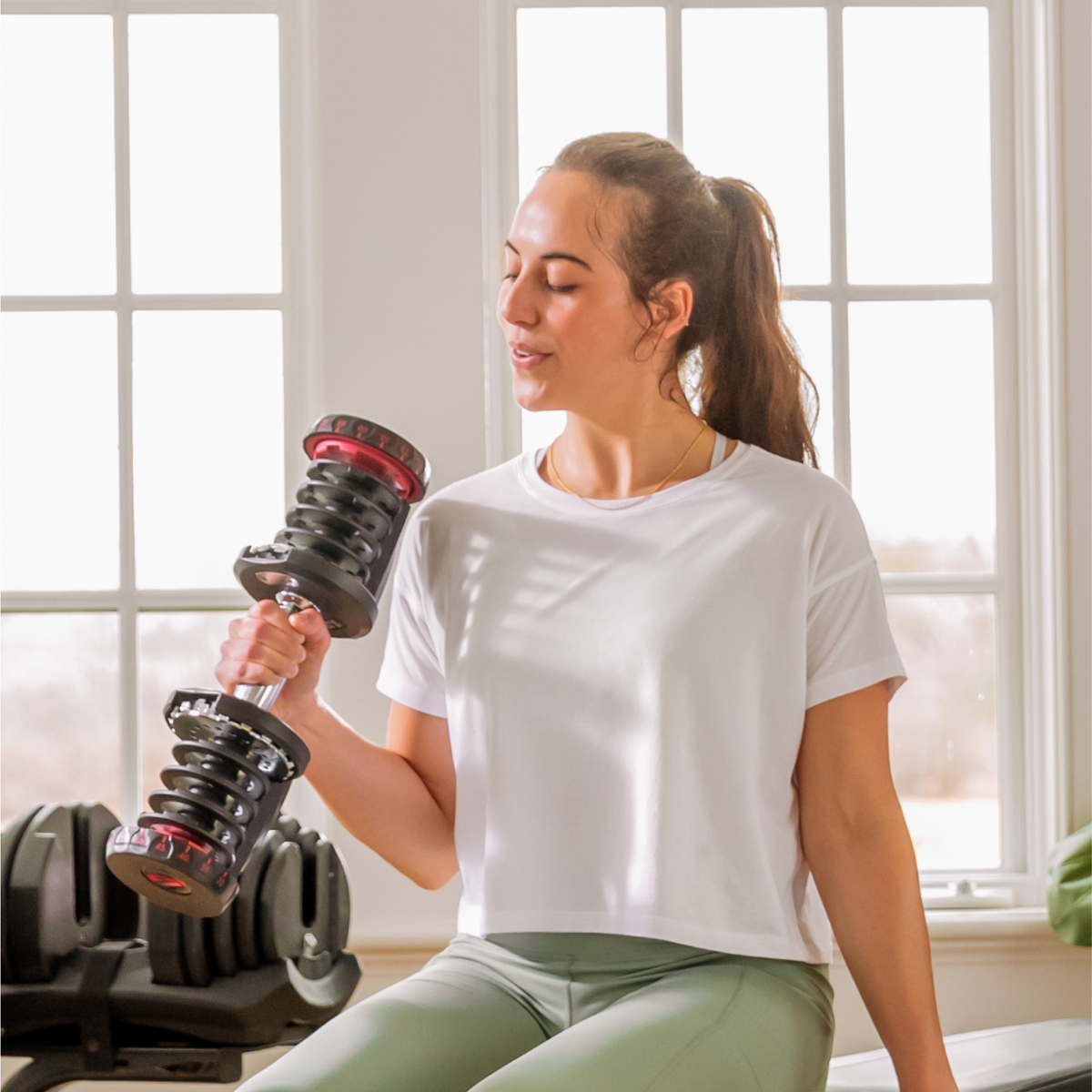 Vrouw die een halter optilt, zittend bij een raam, in een thuissportschool.