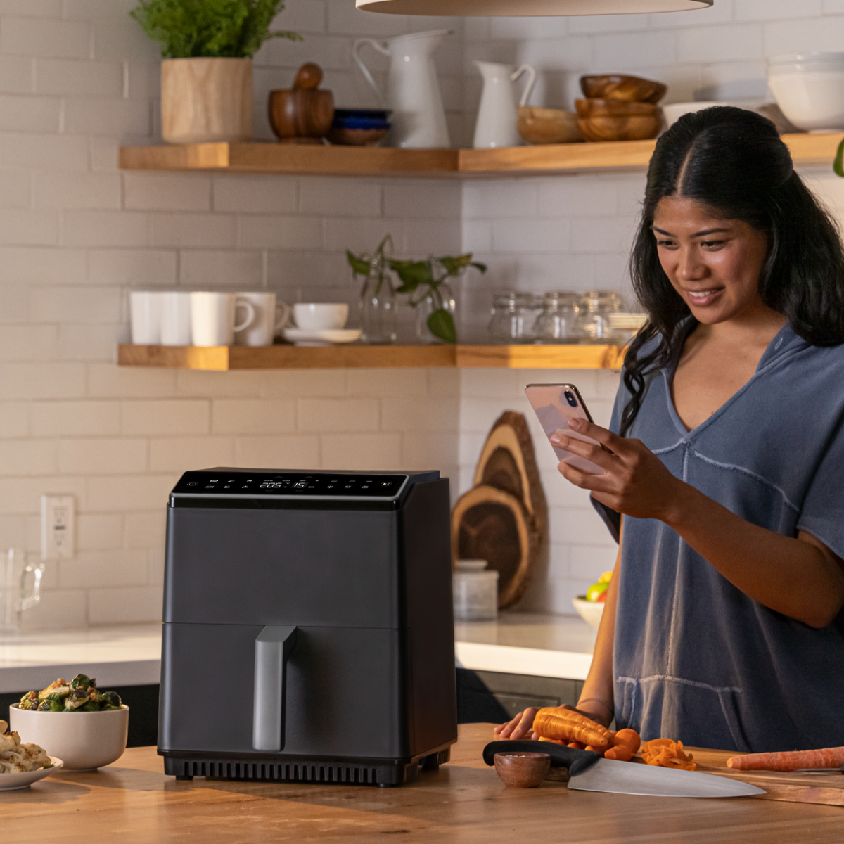 Una donna in cucina guarda il telefono vicino a una friggitrice ad aria e un'area di preparazione del cibo.