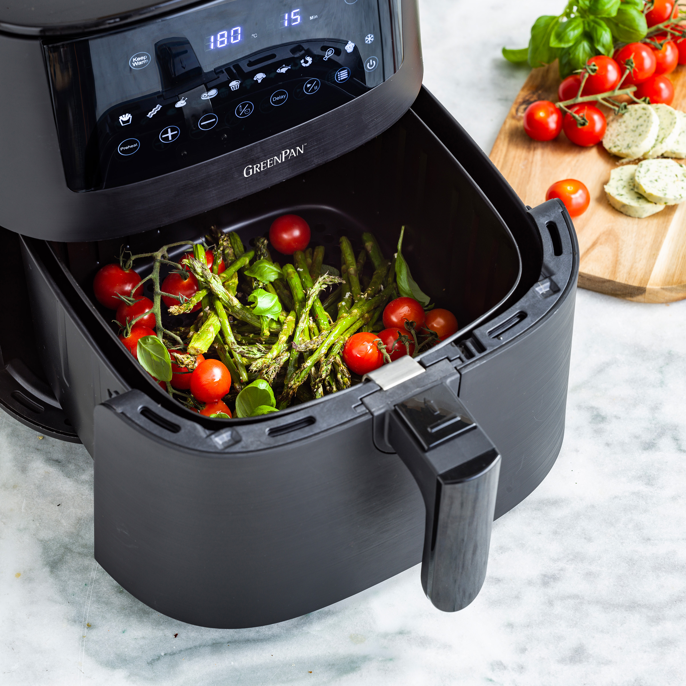 Una friggitrice ad aria nera con pomodori, asparagi e basilico all'interno. Un tagliere di legno accanto.