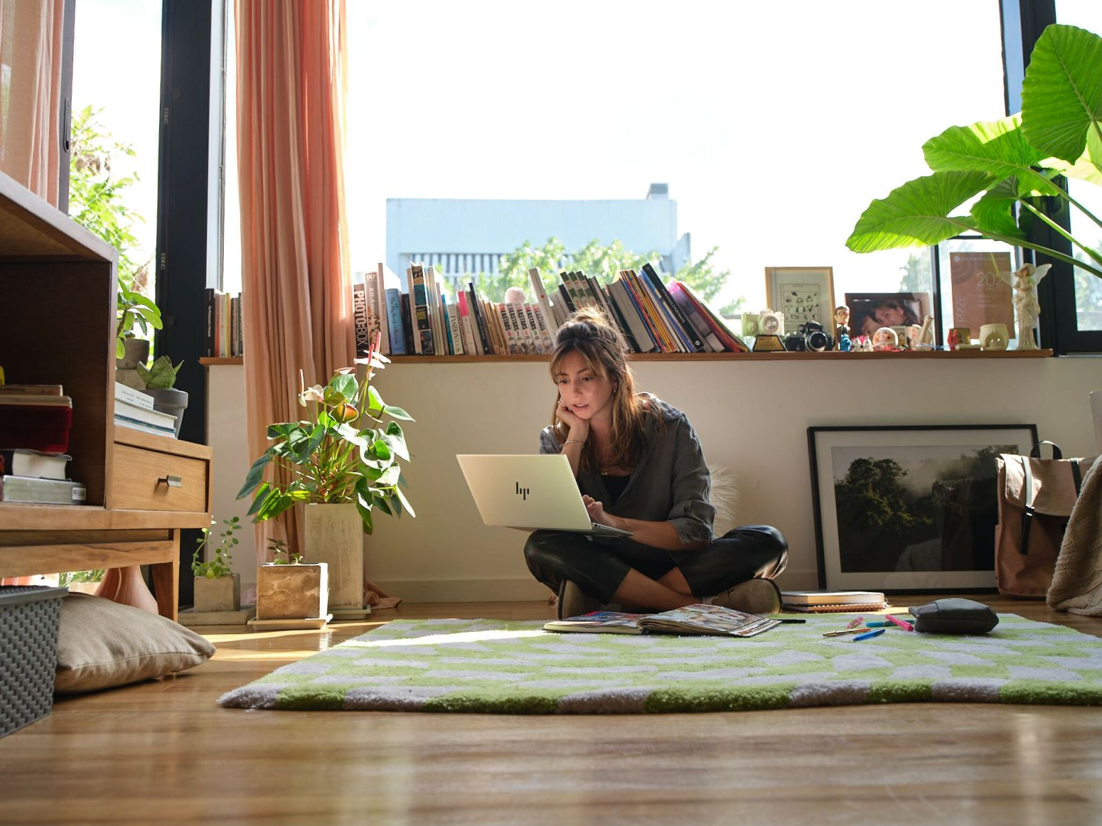 Frau mit Laptop auf Teppich, Fenster, Bücherregal. Grüner Teppich, Holzboden.