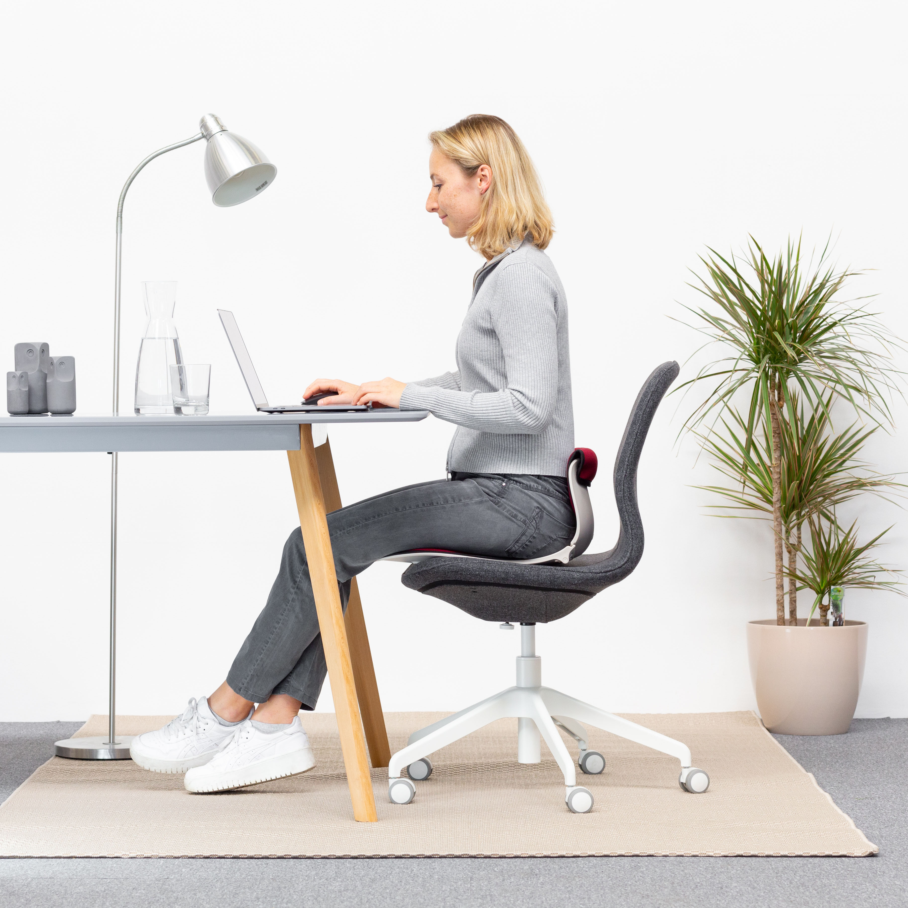 Frau sitzt an einem Schreibtisch und benutzt einen Laptop. Sie trägt ein graues Hemd und eine Hose, mit einem grauen Stuhl.