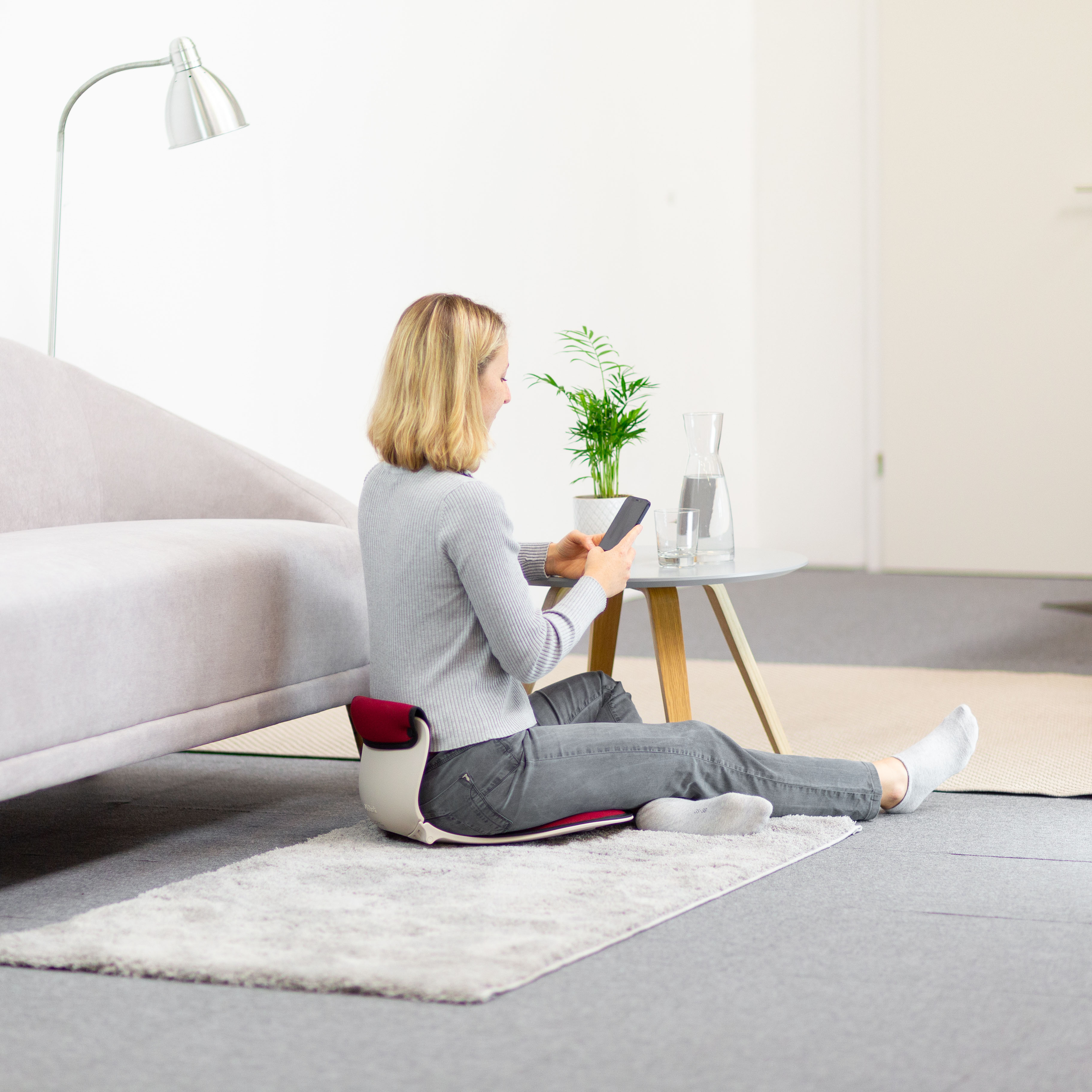 Eine Frau sitzt auf dem Boden und benutzt ein Telefon. Es gibt eine Pflanze und einen Tisch.