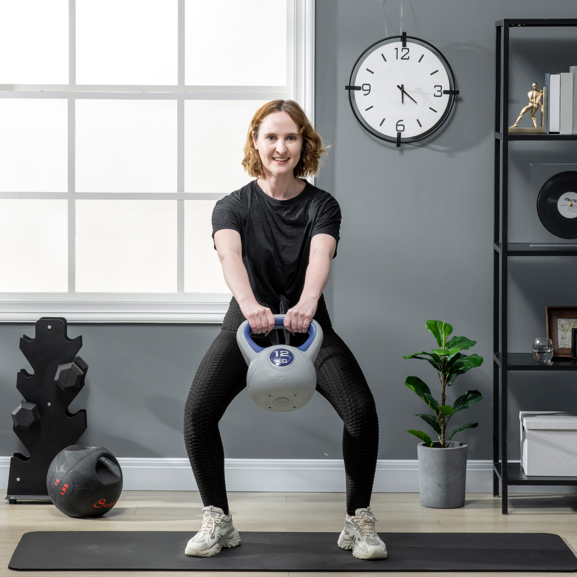 Eine Frau macht Kniebeugen mit einer Kettlebell. Sie befindet sich in einem Home Gym mit Uhr, Pflanze und Gewichten.