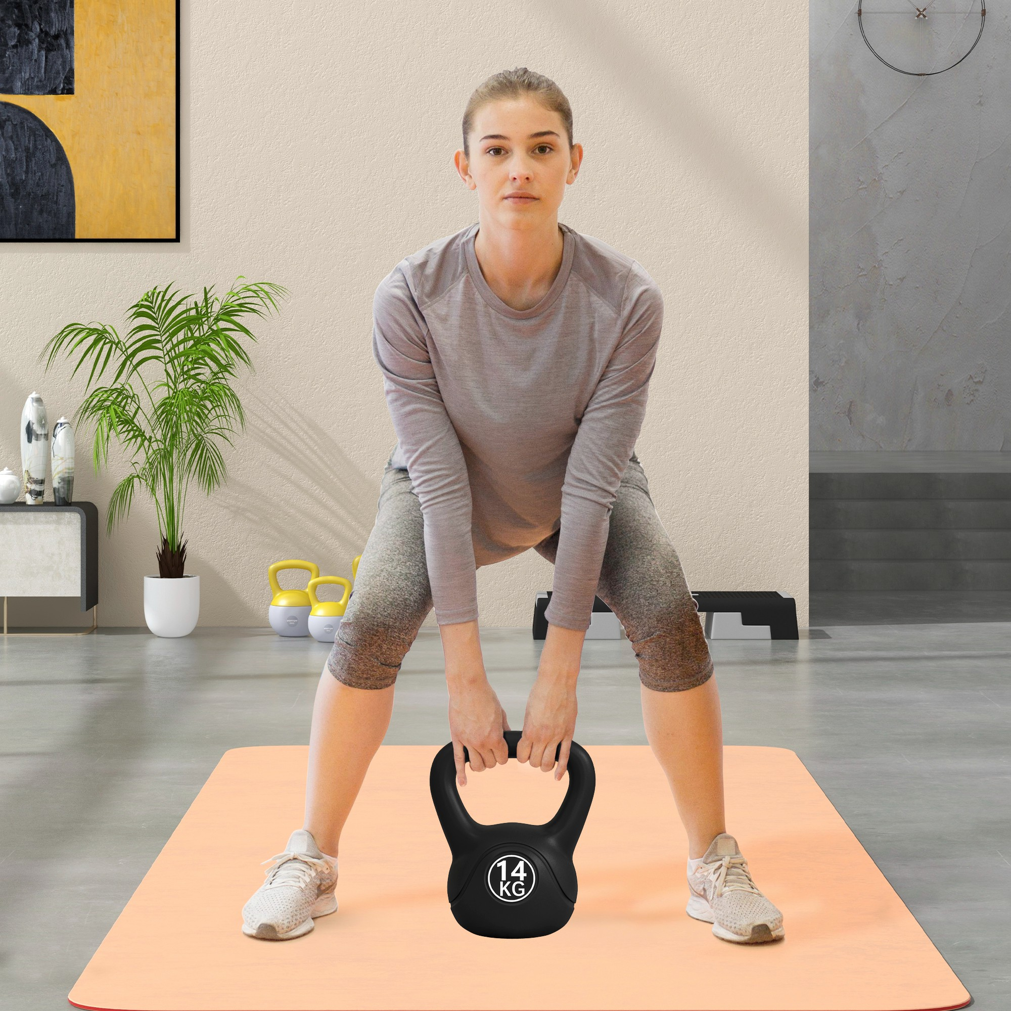 Una mujer en ropa deportiva sosteniendo una pesa rusa negra, de pie sobre una esterilla de yoga naranja.