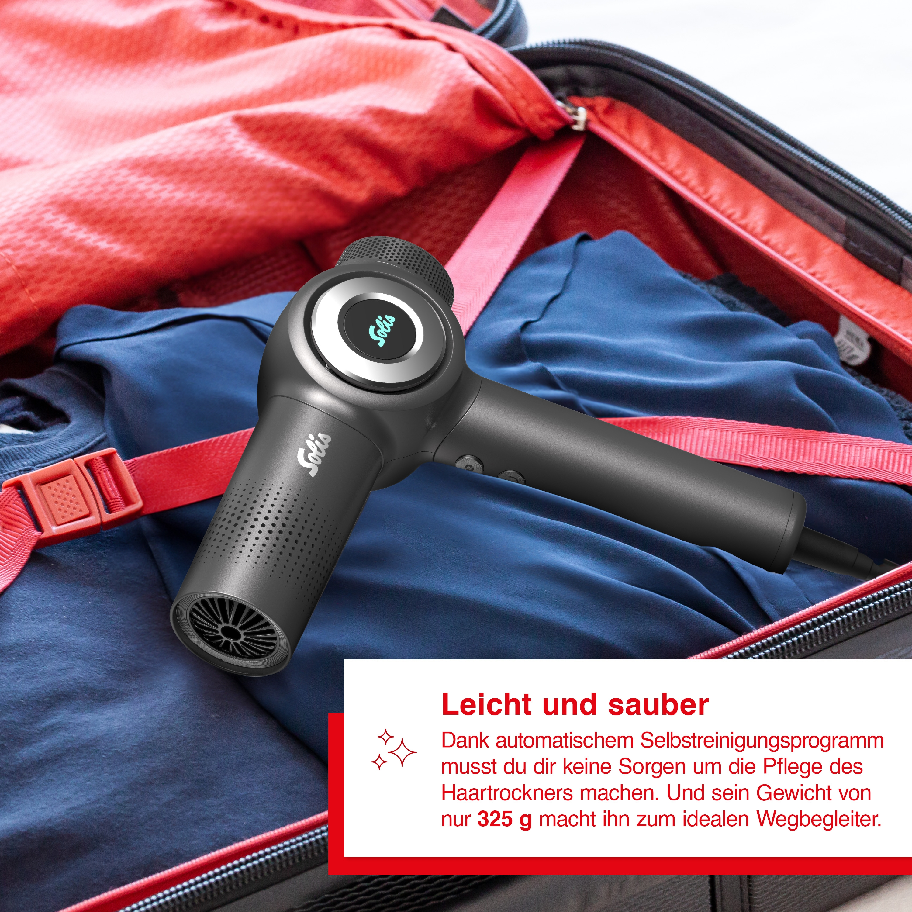 Schwarzer und grauer Fön im roten Koffer. Text auf dem Fön. Offener Koffer mit blauer Kleidung.