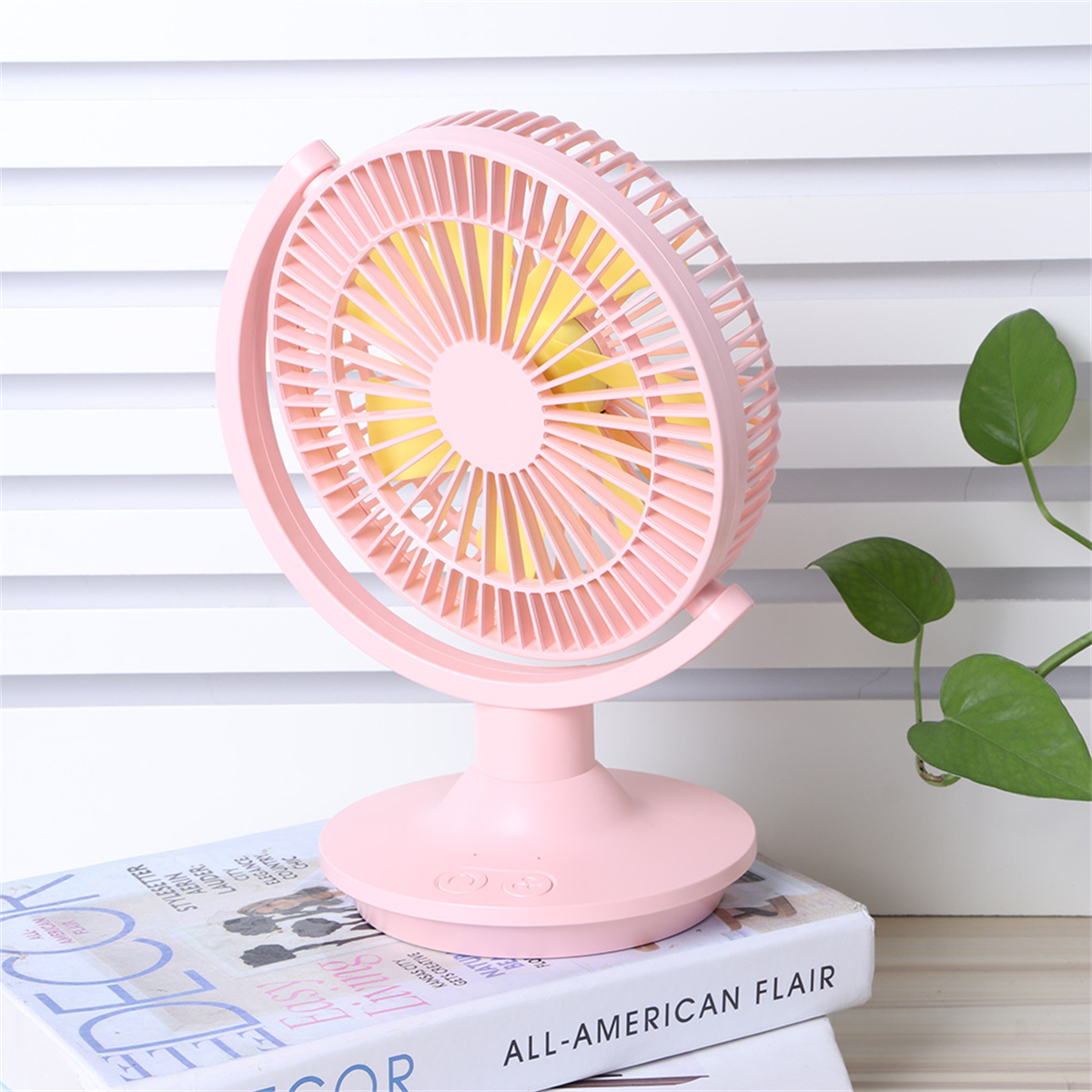 Ein rosa Ventilator mit rundem Kopf und Ständer, auf einem Tisch mit Büchern.