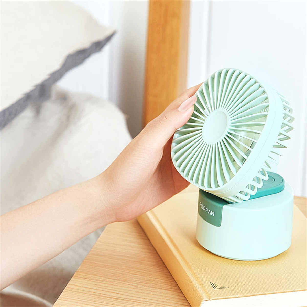 Hand, die einen hellblauen tragbaren Ventilator hält. Der Ventilator steht auf einem türkisfarbenen Sockel, auf einem Buch.