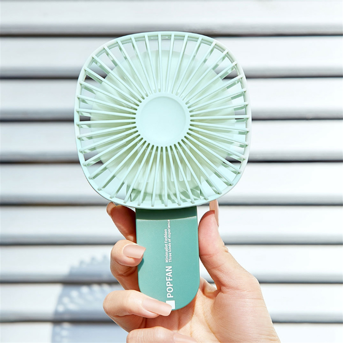 Eine Hand hält einen mintgrünen Ventilator, im Hintergrund weiße Jalousien.