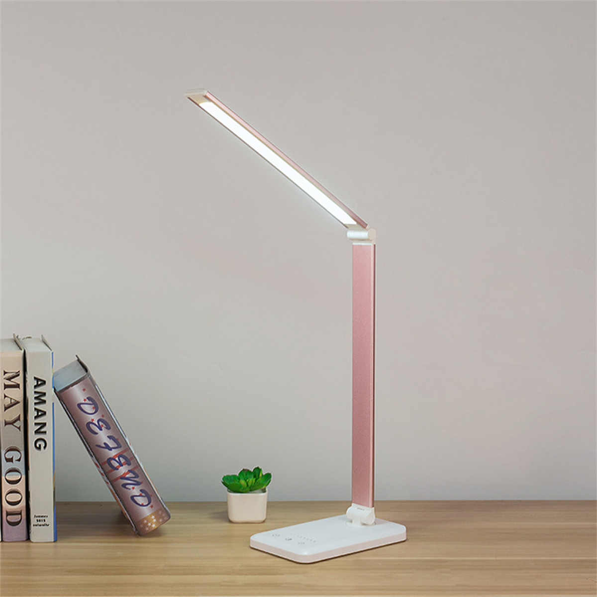 Eine moderne Schreibtischlampe mit pinkfarbenem Stiel und weißem Sockel, Bücher und Pflanze.