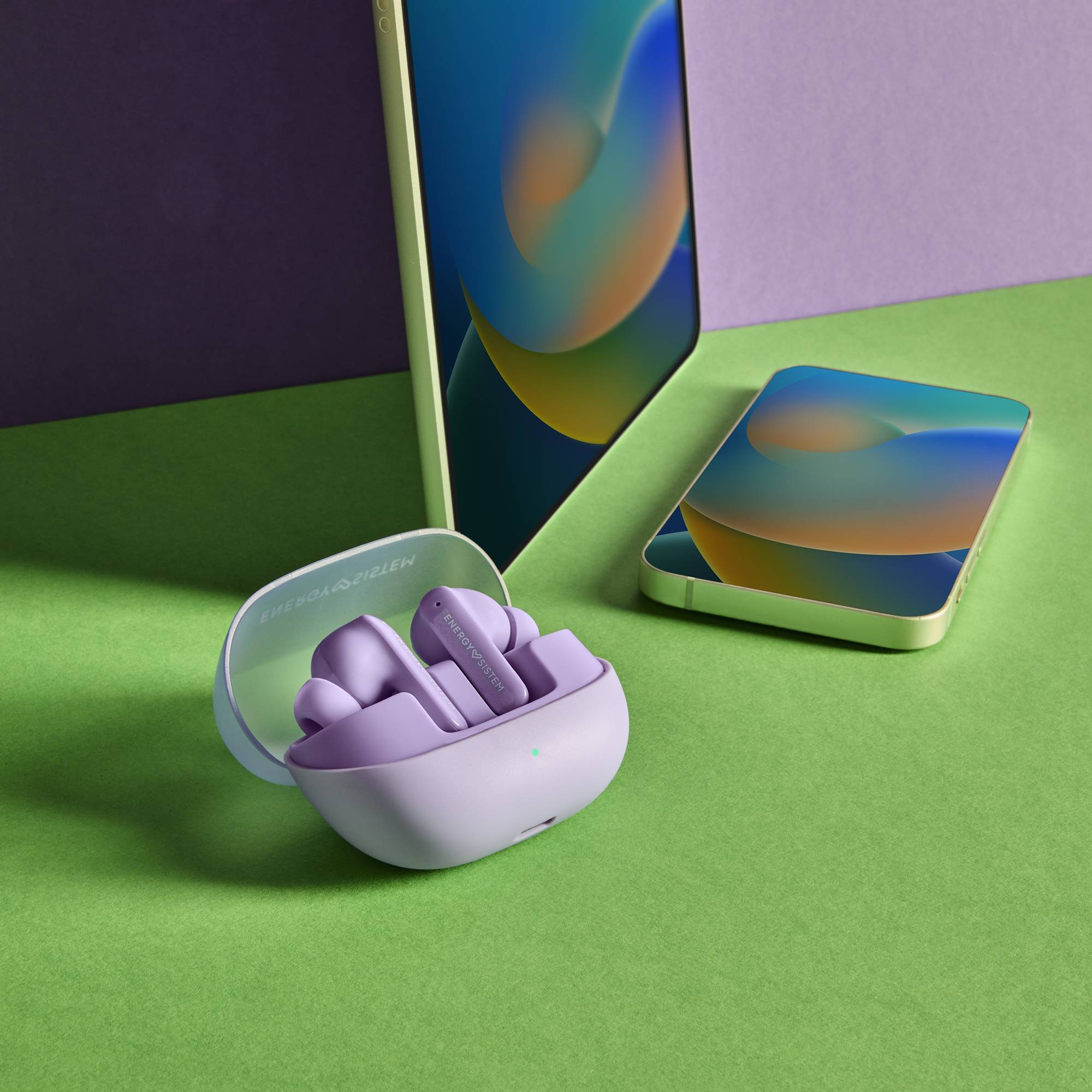 Paarse oortelefoons in een hoesje, naast een telefoon en tablet op groene en paarse oppervlakken.