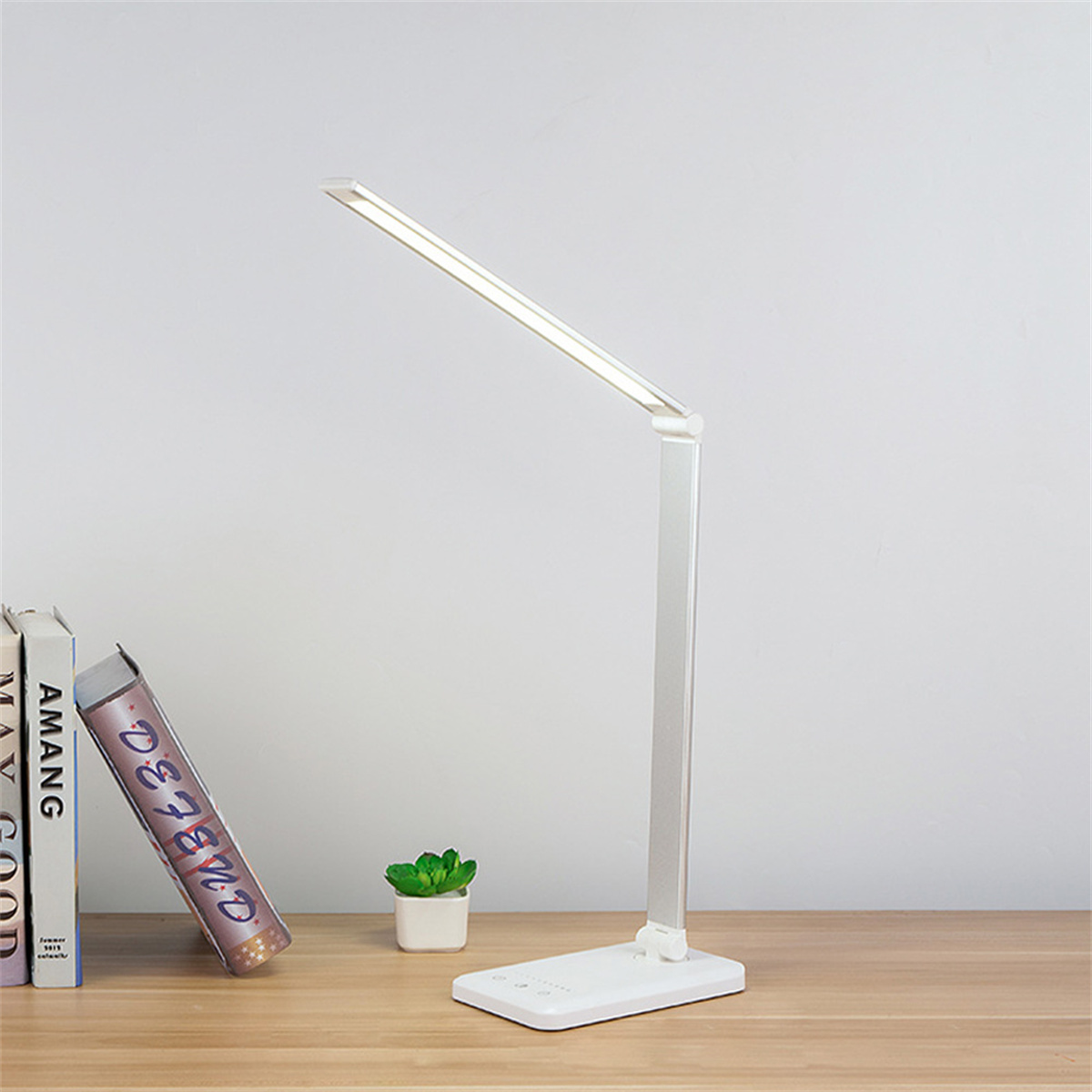 Eine moderne silberne Schreibtischlampe mit hellem Licht, neben Büchern und einer kleinen Pflanze auf einem hölzernen Schreibtisch.