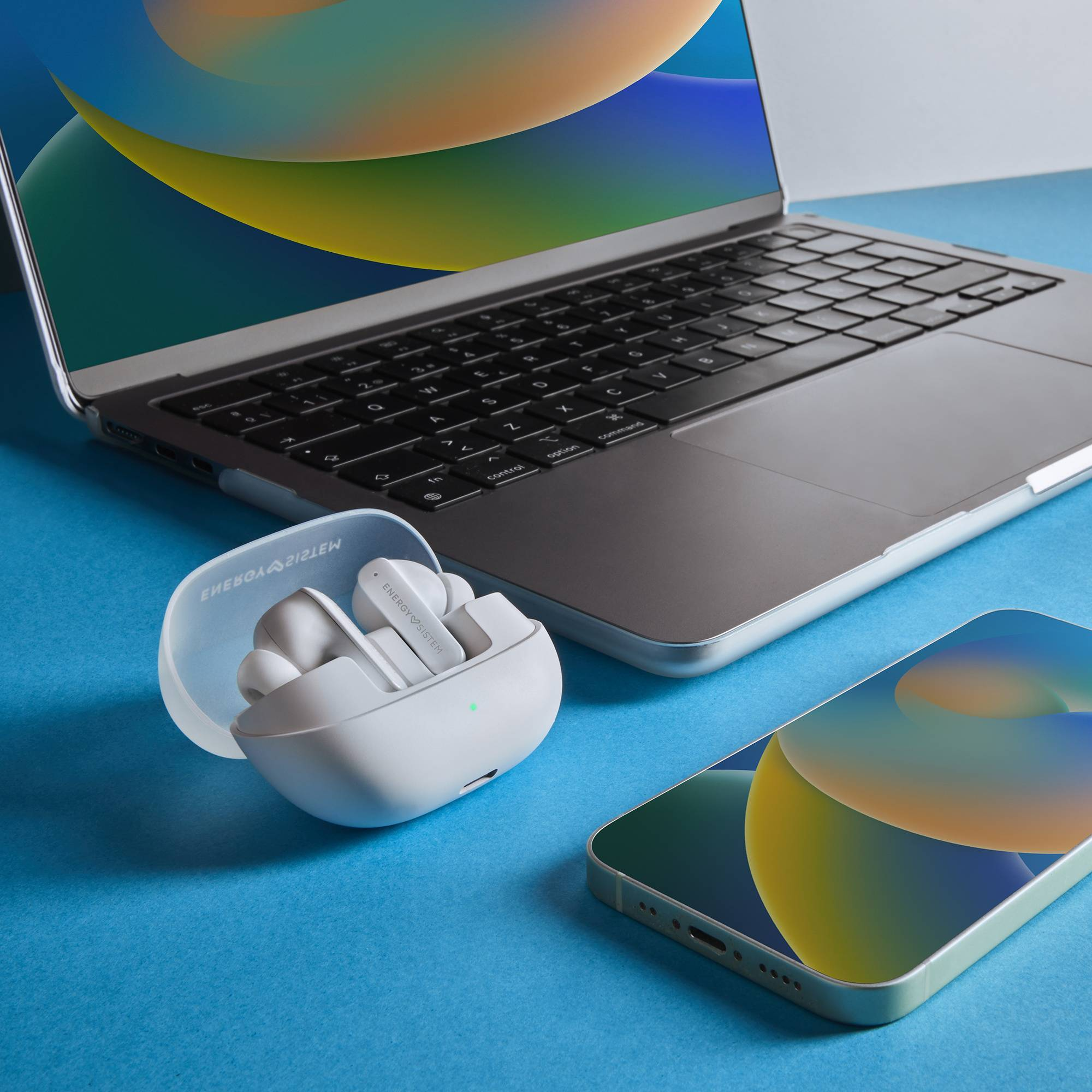 Kabellose Ohrhörer in einem Ladecase, neben einem Laptop und einem Smartphone auf blauer Oberfläche.