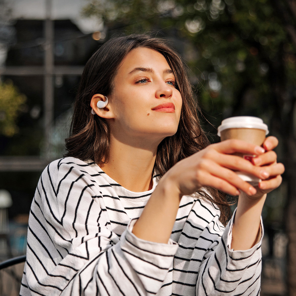 Donna con auricolari, che tiene un caffè, guarda in su, con una camicia a righe.