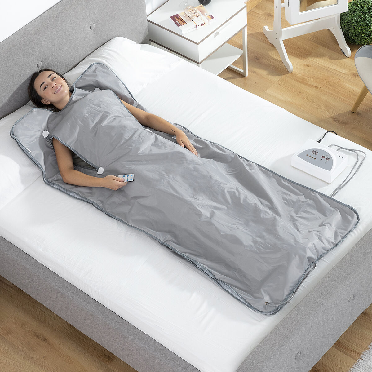 Una donna sdraiata su un letto coperta da una coperta per sauna a infrarossi grigia, con un pannello di controllo.