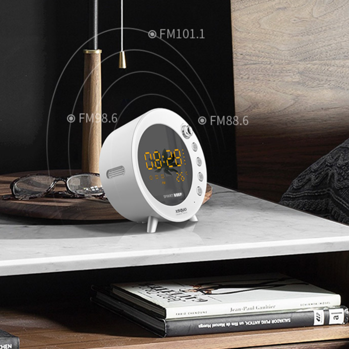 Despertador blanco. Tiene una pantalla digital y está sobre una mesita de noche con libros.