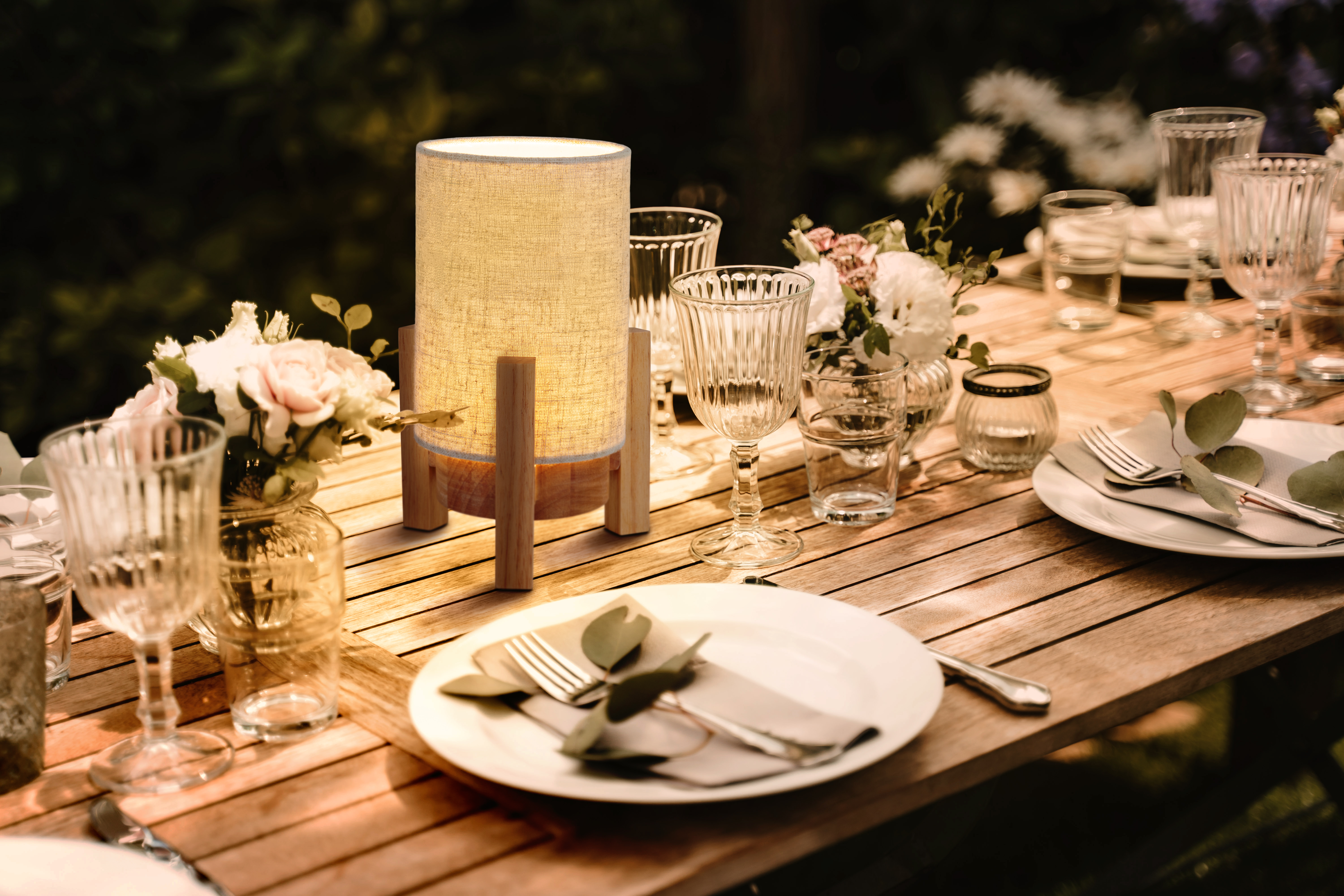 Eine Tischdekoration mit Lampe, Tellern und Blumen, in einer Außenumgebung mit Sonnenlicht.