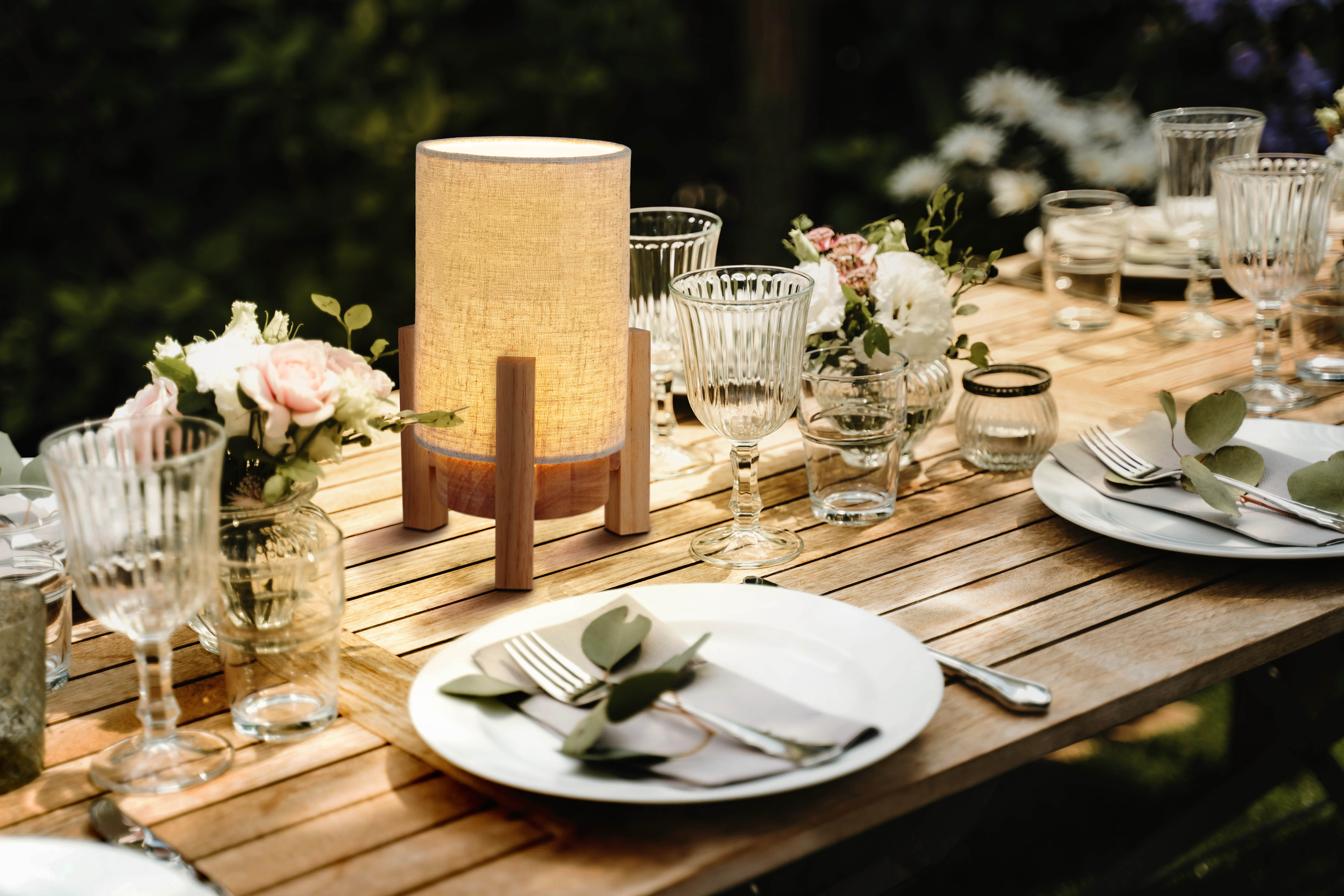 Tisch im Freien gedeckt. Tisch mit Lampe, Gläsern, Tellern und Blumen. Sonniger Tag.