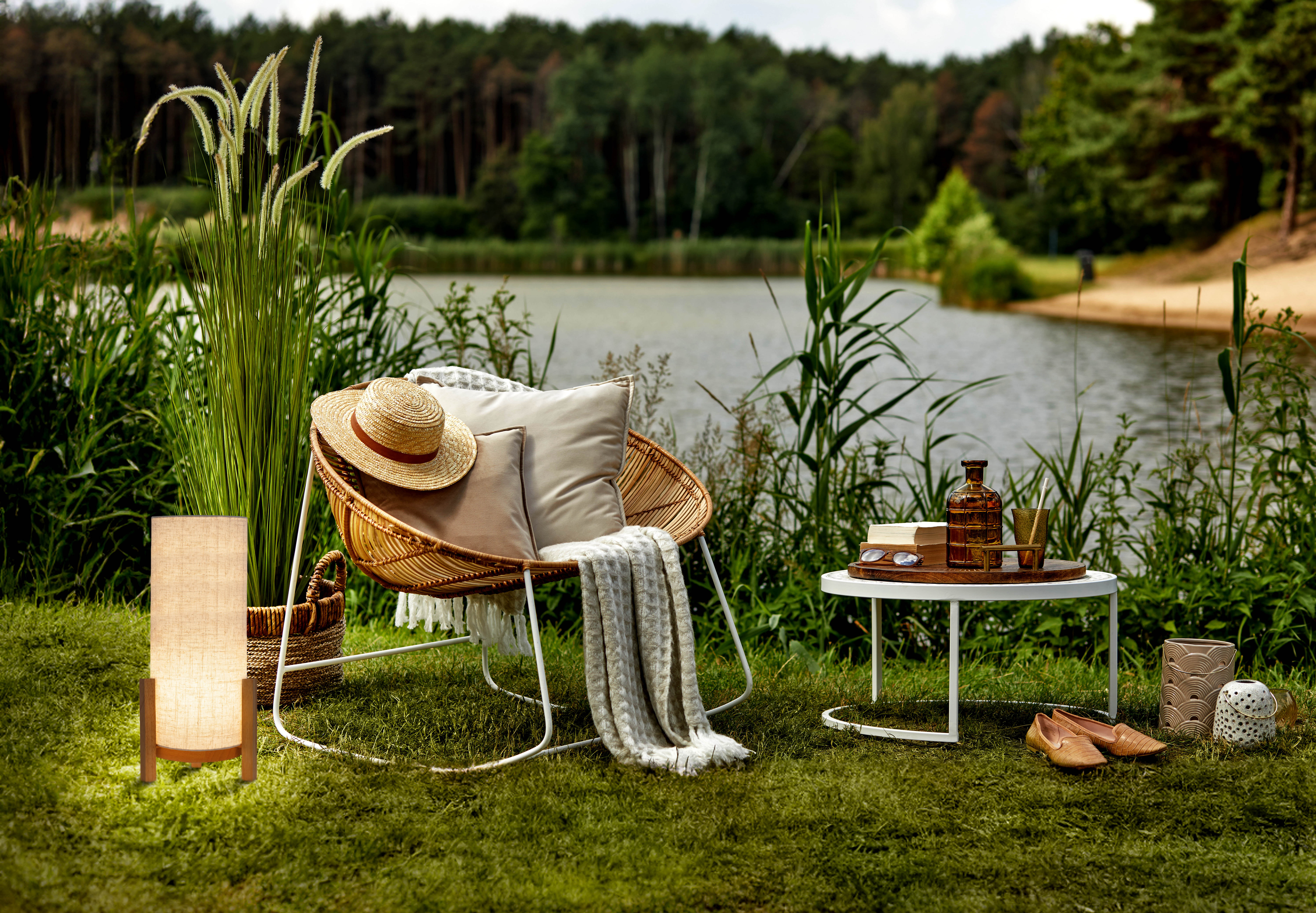 Szene im Freien mit Stuhl, Lampe, Tisch und See im Hintergrund. Gras und Bäume.