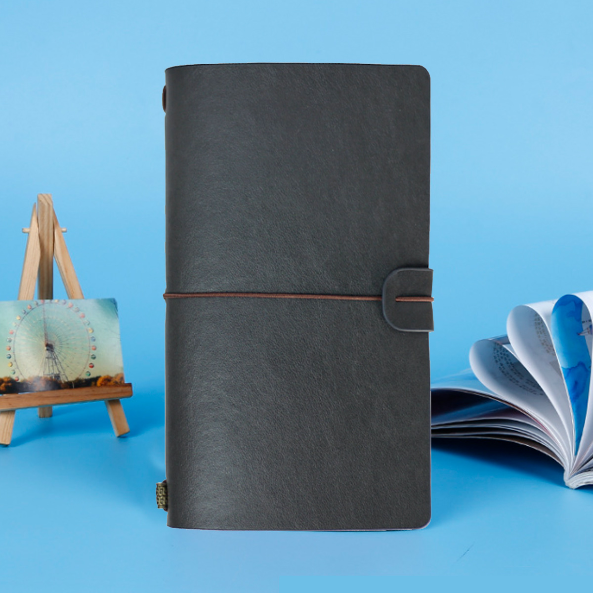 Ein dunkelgraues Lederjournal mit braunem Riemen. Eine kleine Staffelei und eine Zeitschrift im Hintergrund.