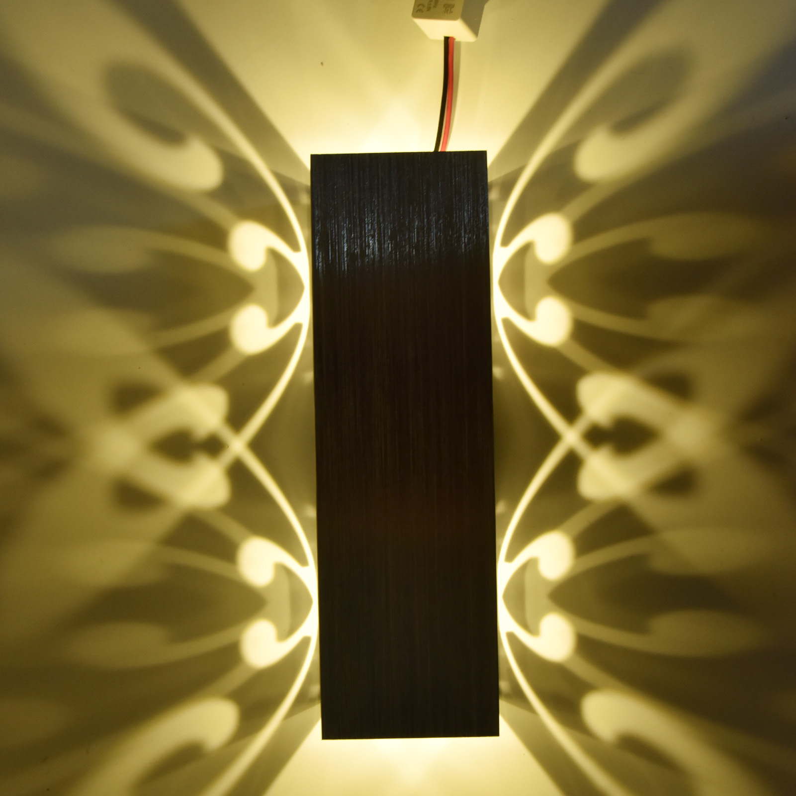 Wandleuchte mit zentralem dunklem Paneel und Licht, das symmetrische Muster an die Wand wirft.