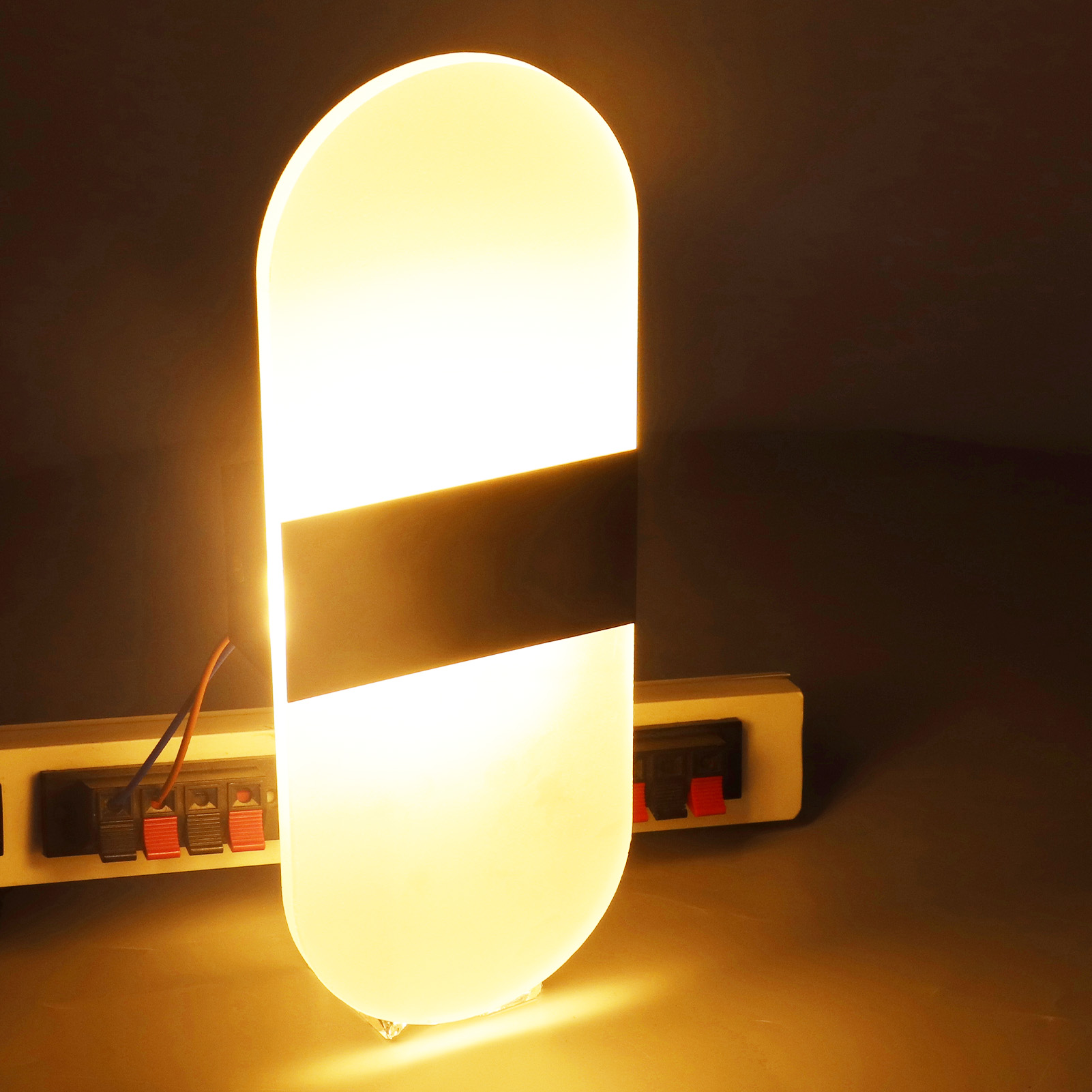 Eine hohe, weiß-schwarze LED-Lampe mit schwarzem Streifen. Sie steht in einem dunklen Raum, eine Steckdose ist in der Nähe.