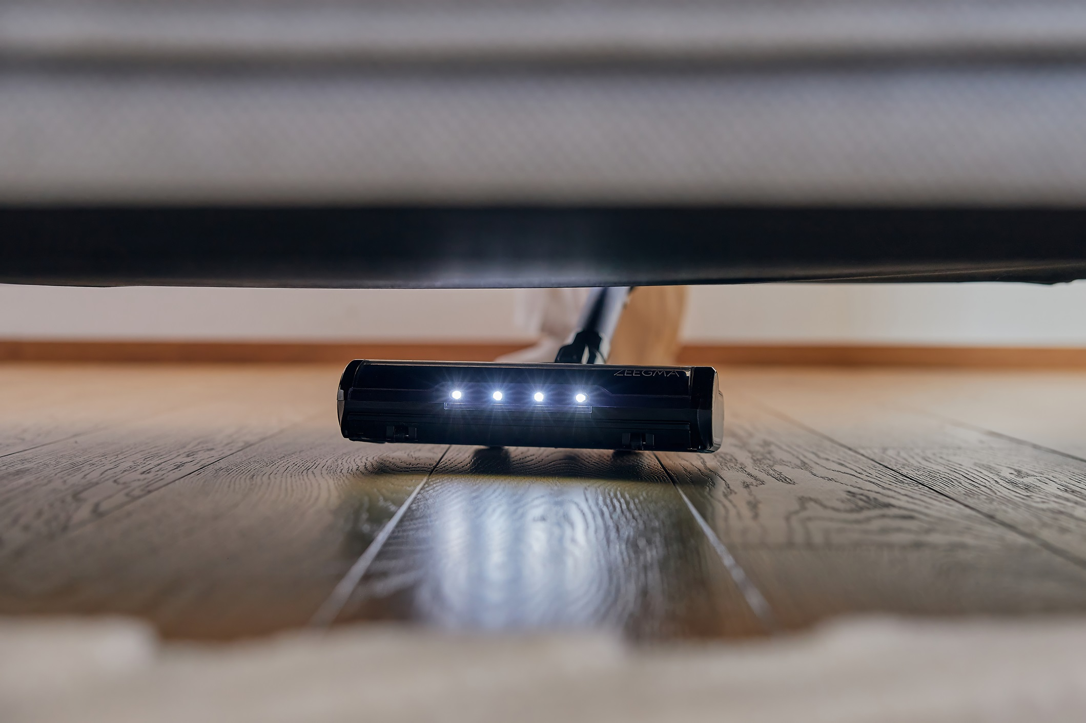 Schwarzer Staubsaugerkopf unter einem Bett. Der Kopf hat 4 Lichter und befindet sich auf einem Holzboden.