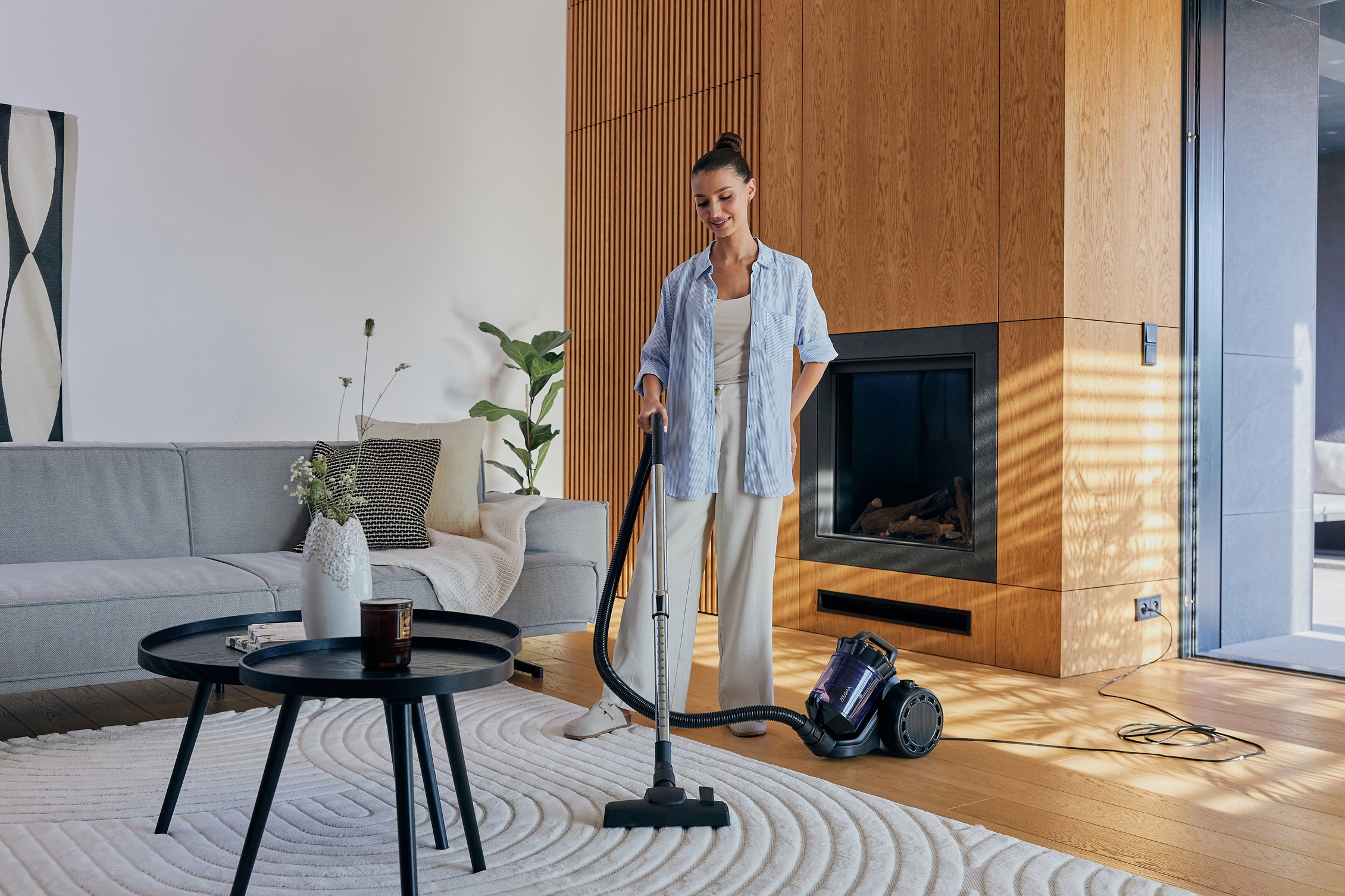 Kobieta odkurza dywan w salonie. Jest sofa, stoliki kawowe i kominek.