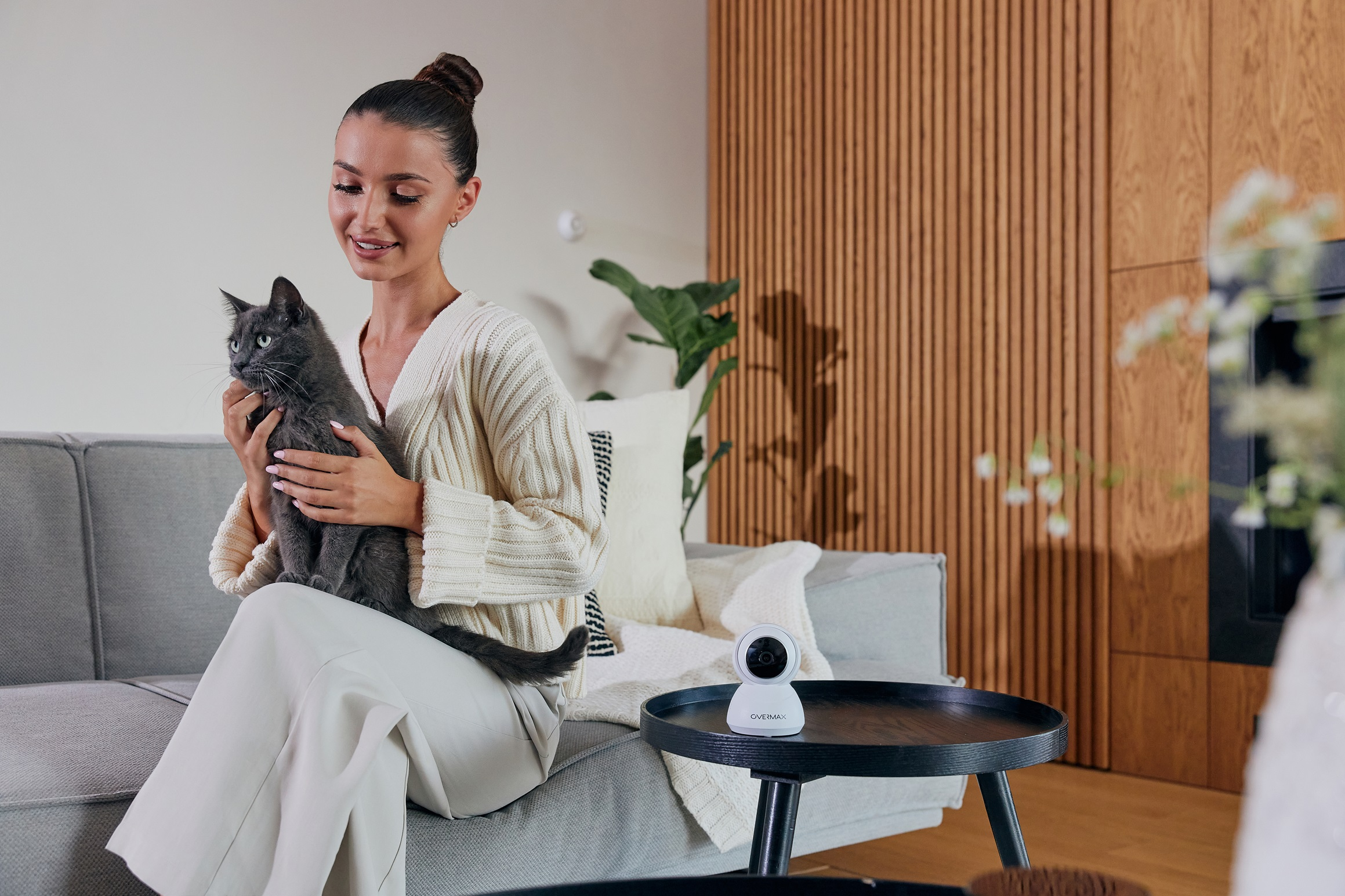 Kobieta głaszcze szarego kota, siedząc na sofie, z kamerą bezpieczeństwa w pobliżu.