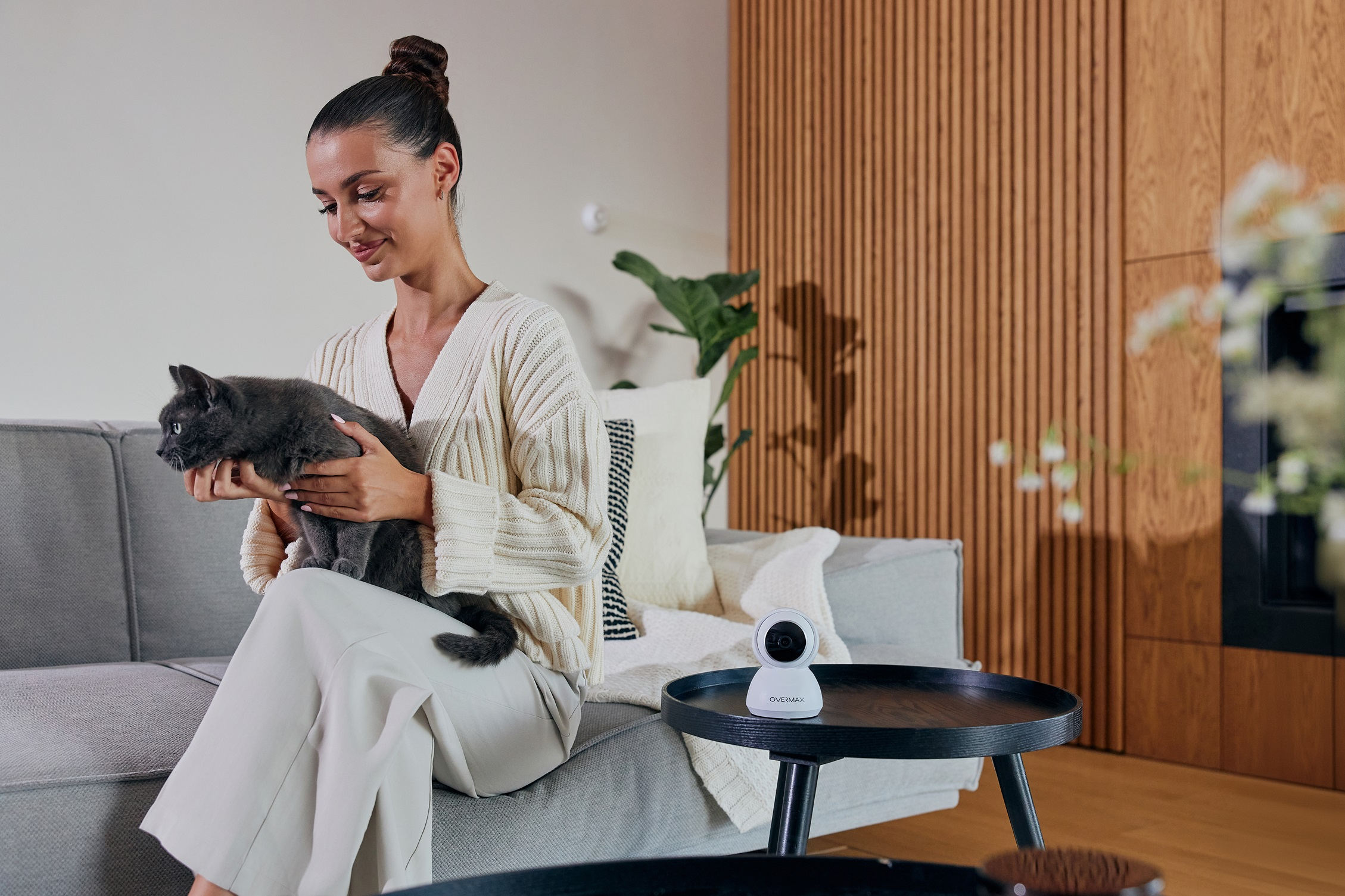 Kobieta trzymająca szarego kota na sofie obok kamery bezpieczeństwa.