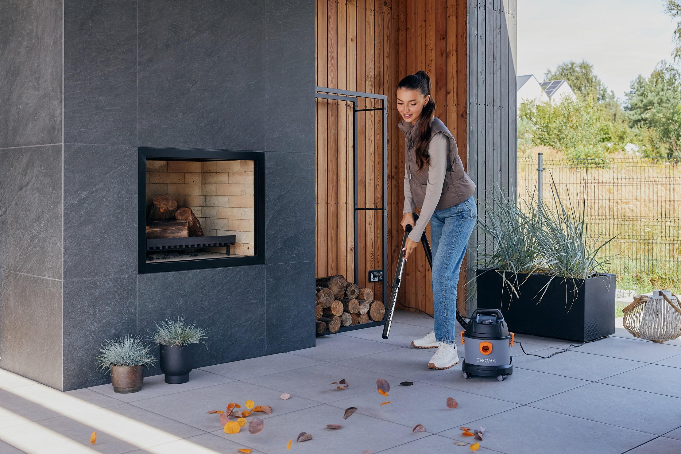 Kobieta odkurza liście na patio. Kominek i panele drewniane w tle, odkurzacz na pierwszym planie.