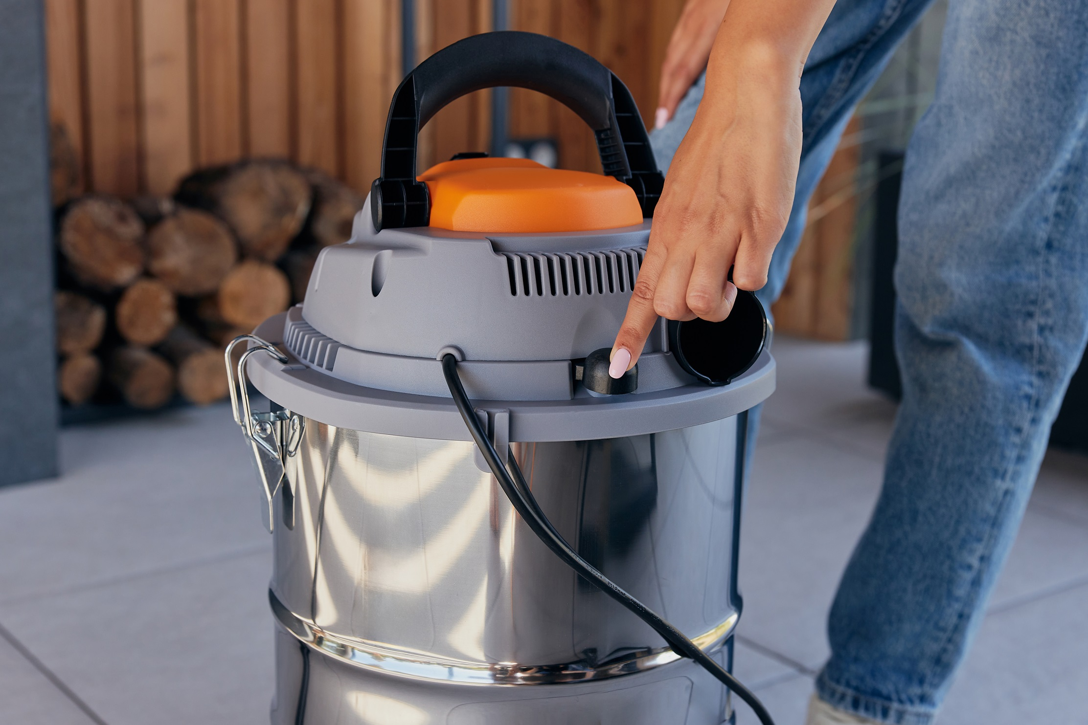 Ręka osoby naciskająca przycisk na srebrnym i pomarańczowym odkurzaczu. W tle drewno i podłoga.