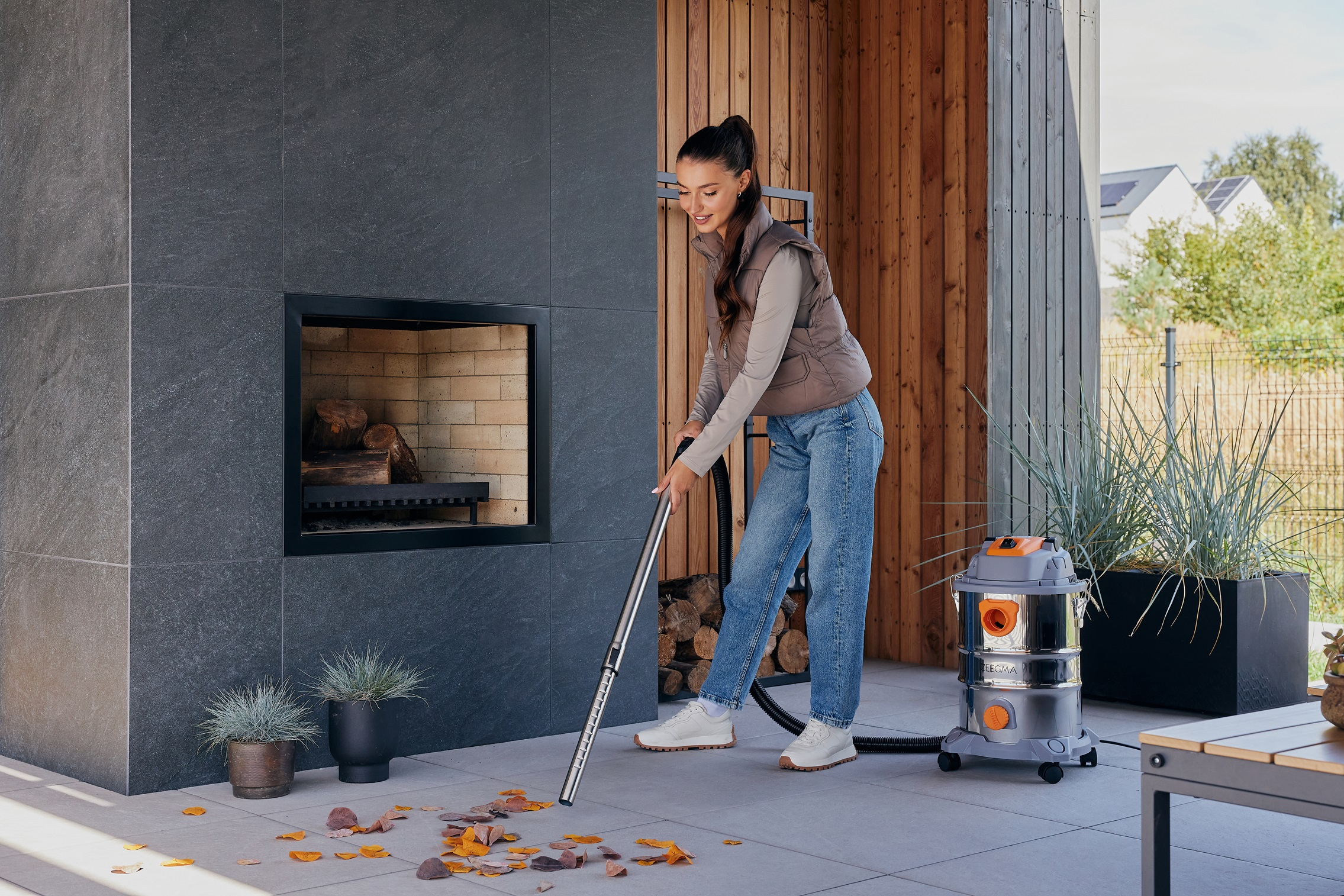 Kobieta odkurzająca patio, liście na ziemi. Szara kamienna ściana w tle.