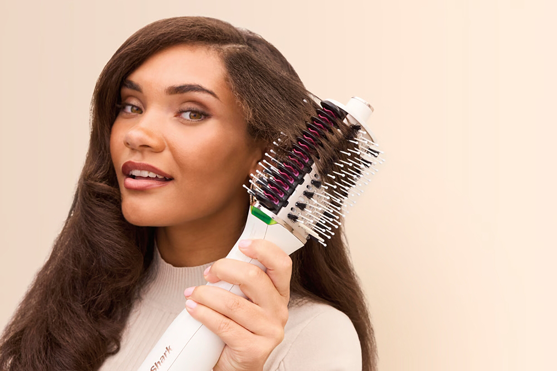 Kobieta z falowanymi włosami używa szczotki do suszenia włosów. Uśmiecha się i trzyma szczotkę w stronę kamery.