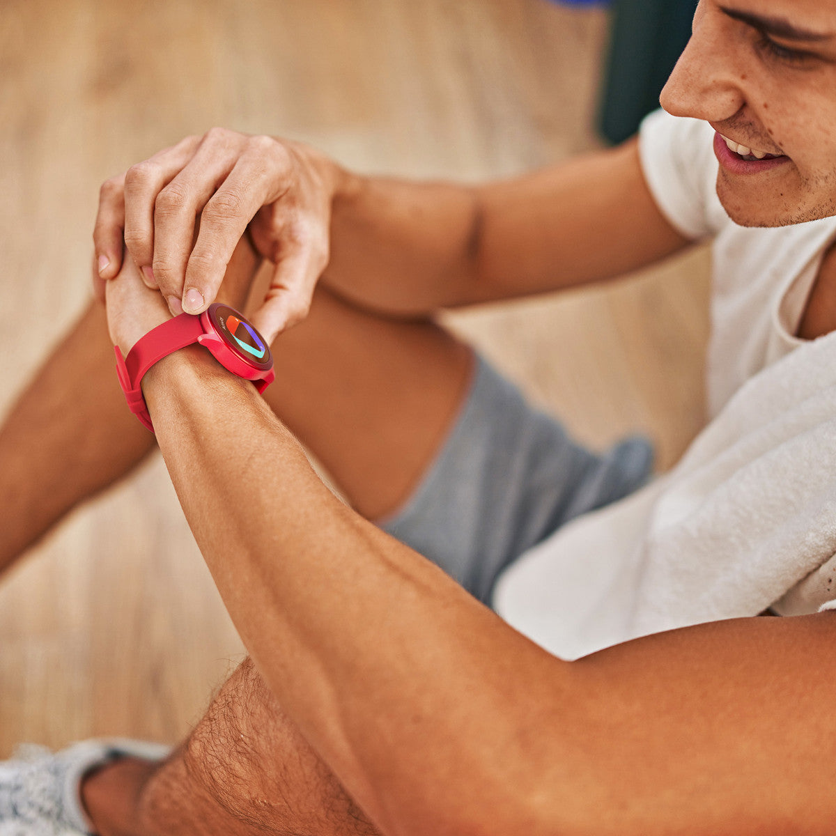 Persona con smartwatch rosso, tocca lo schermo. Pavimento in legno. Viso sorridente. Luce naturale.