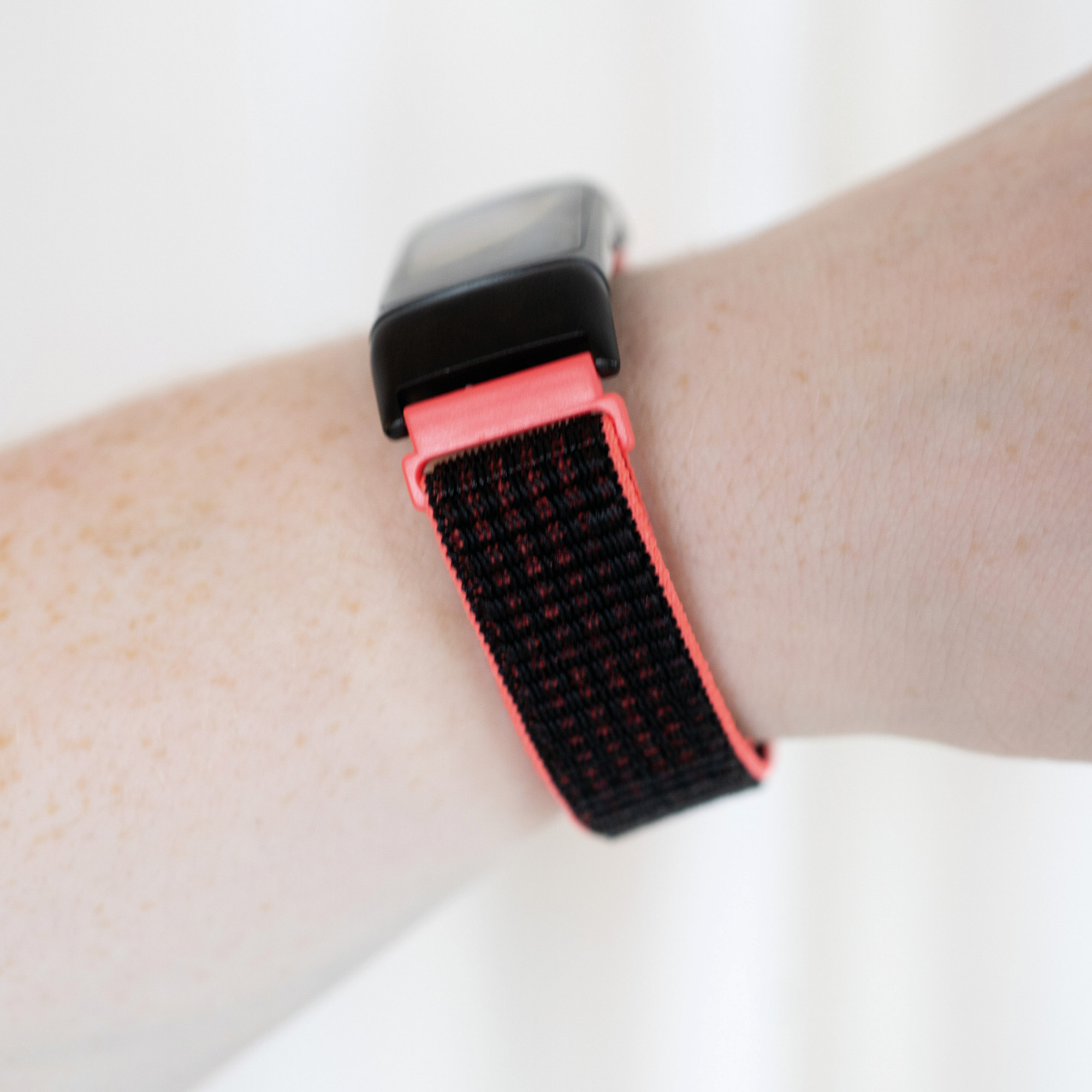 Arm einer Person mit einer Smartwatch und einem korallenfarbenen und schwarzen Armband. Der Arm hat Sommersprossen.