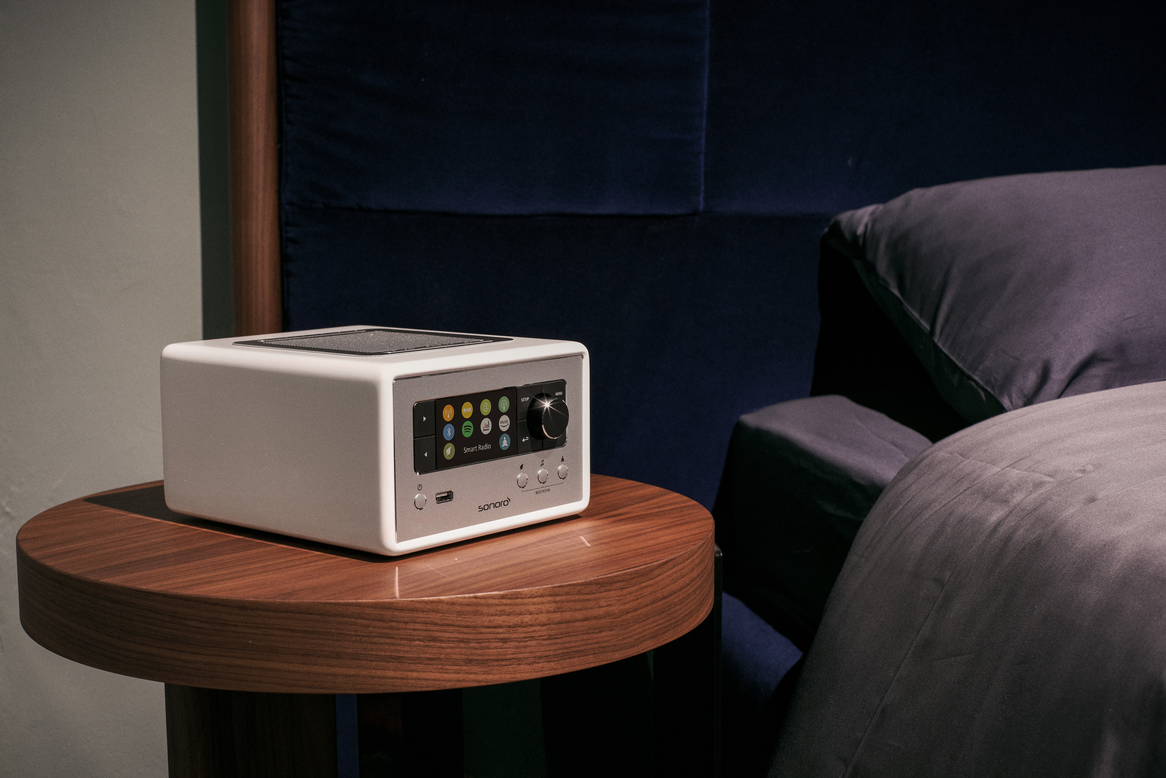 Silver smart radio on a dark wood table. Bed in the background.