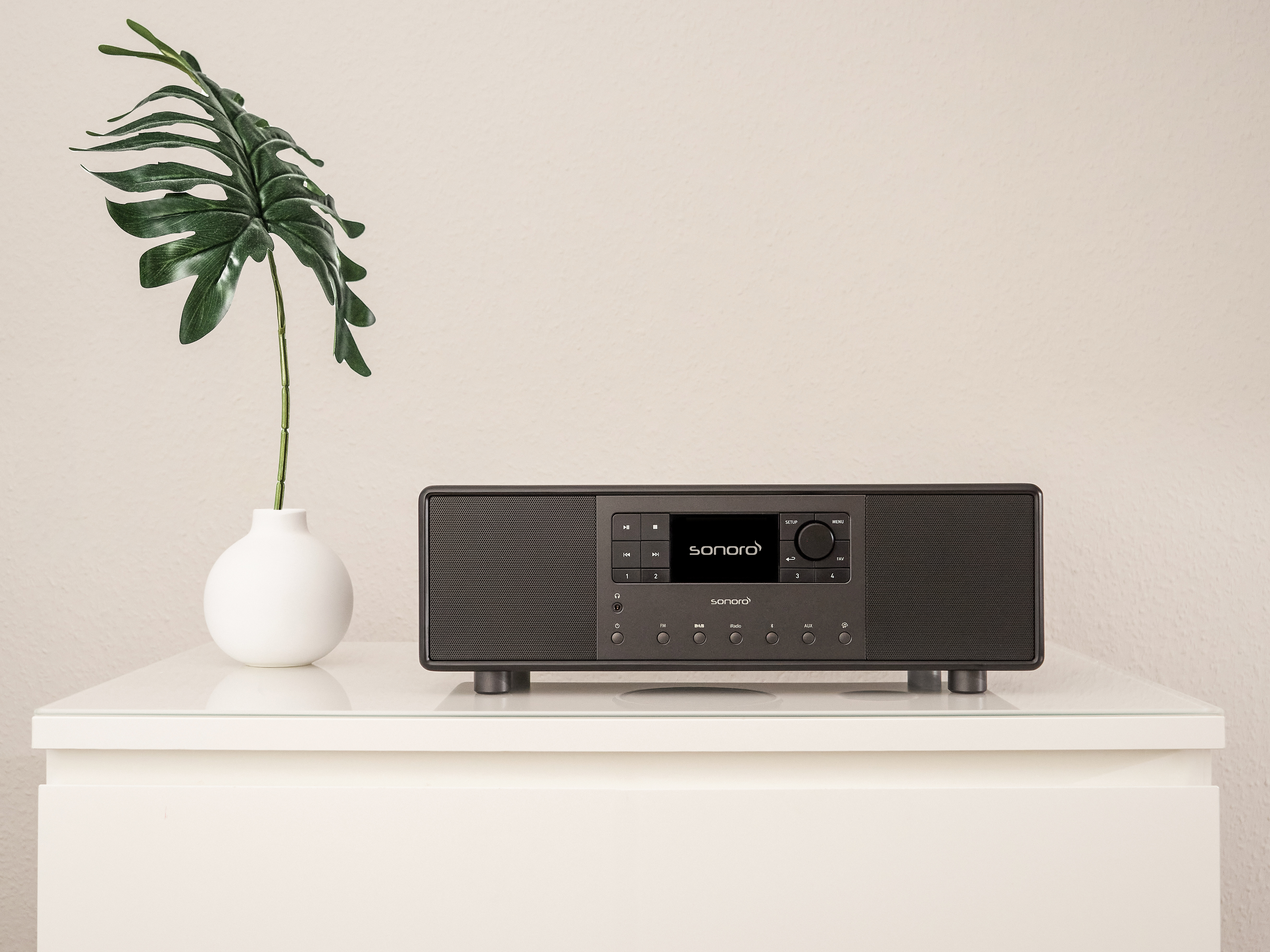 Zwarte Sonoro radio op witte tafel, witte vaas met plant. Effen witte muur.